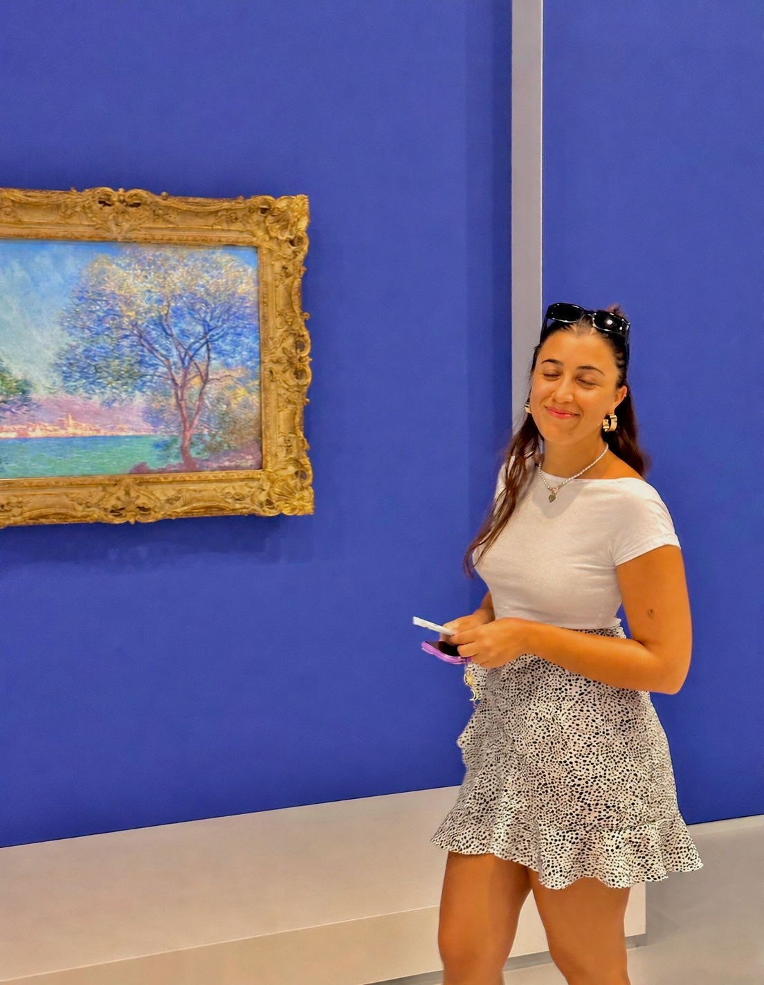 A woman with sunglasses on her head, wearing a white t-shirt and a short, ruffled, animal print skirt, standing in an art gallery in front of a framed colorful landscape painting, smiling with her eyes closed and holding a phone and a small object in her hands.