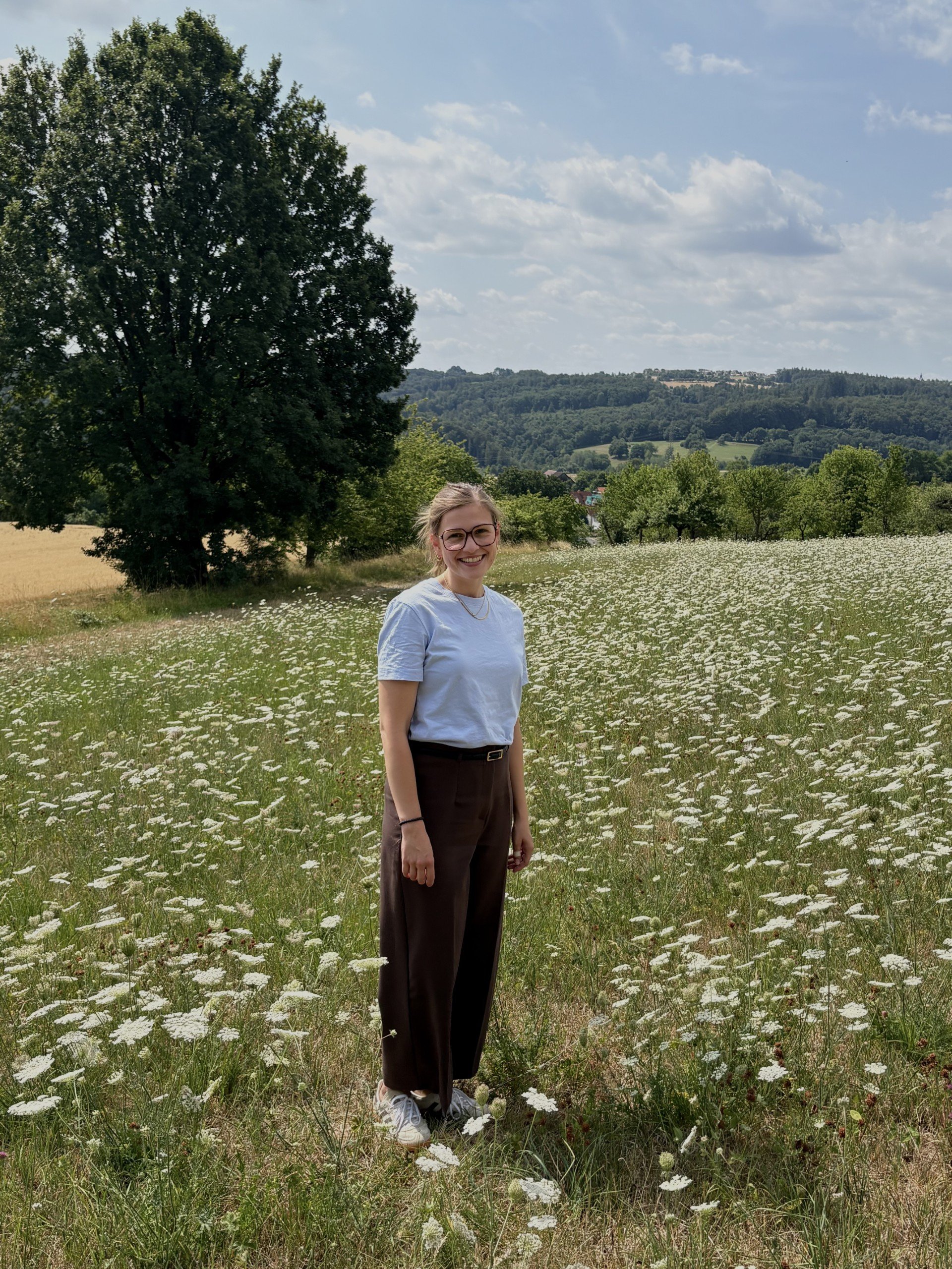 Eine lachende junge Frau mit Brille steht in einem Feld voller weißer Blumen, im Hintergrund Bäume und ein Hügel mit Häusern unter einem bewölkten Himmel.