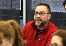 A man with glasses and a beard wearing a red shirt appears to be speaking in a meeting or discussion.