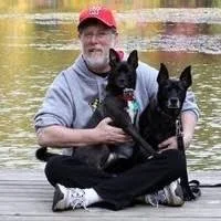 An older man sitting outdoors on a wooden surface, holding two black dogs, with a body of water in the background.