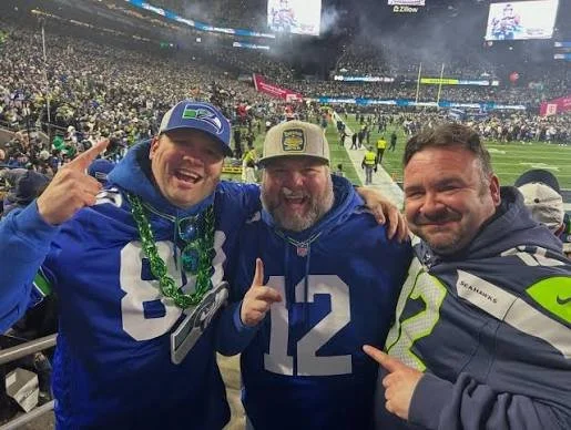 Three men in Seahawks and NFL jerseys at a stadium, smiling and pointing, with a large crowd and field in the background.