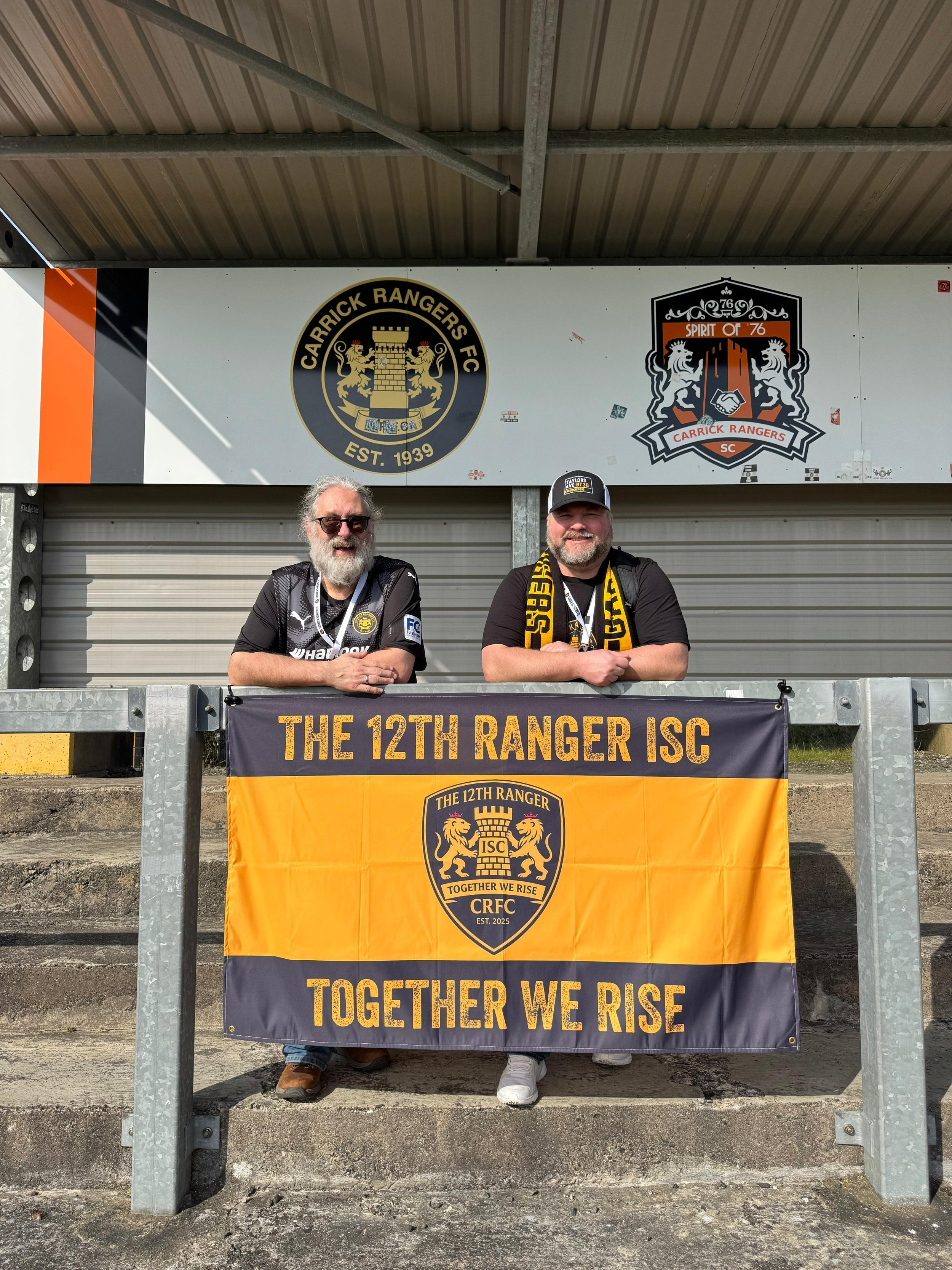Two men behind a banner reading "The 12th Ranger ISC," with additional text "Together We Rise." The men are wearing ranger jerseys and scarves, standing in front of a metal barrier and signs for Carrick Rangers FC.