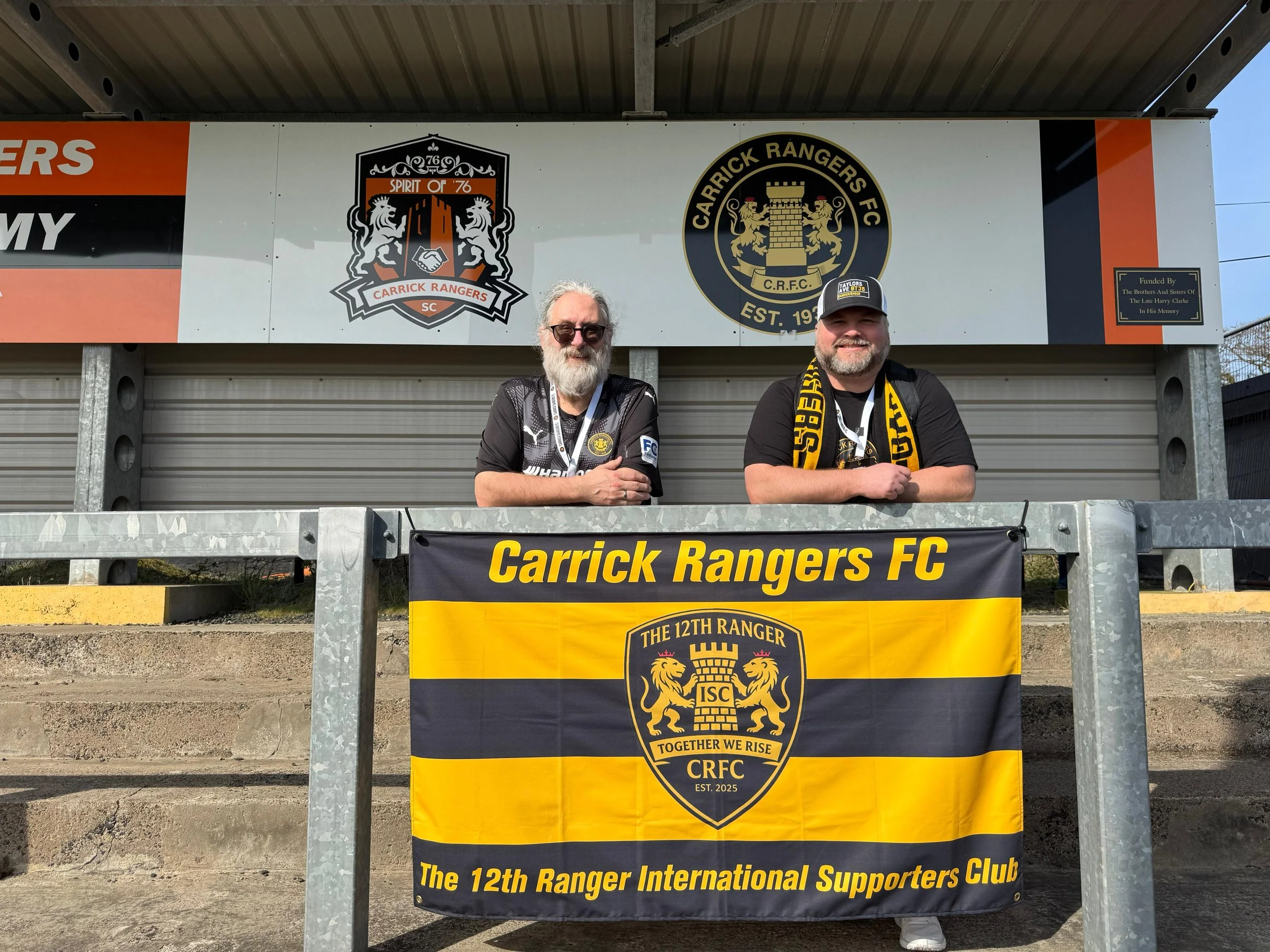 Two men standing in front of sports team banners for Carrick Rangers FC, with logos of the team and crest, smiling outdoors.