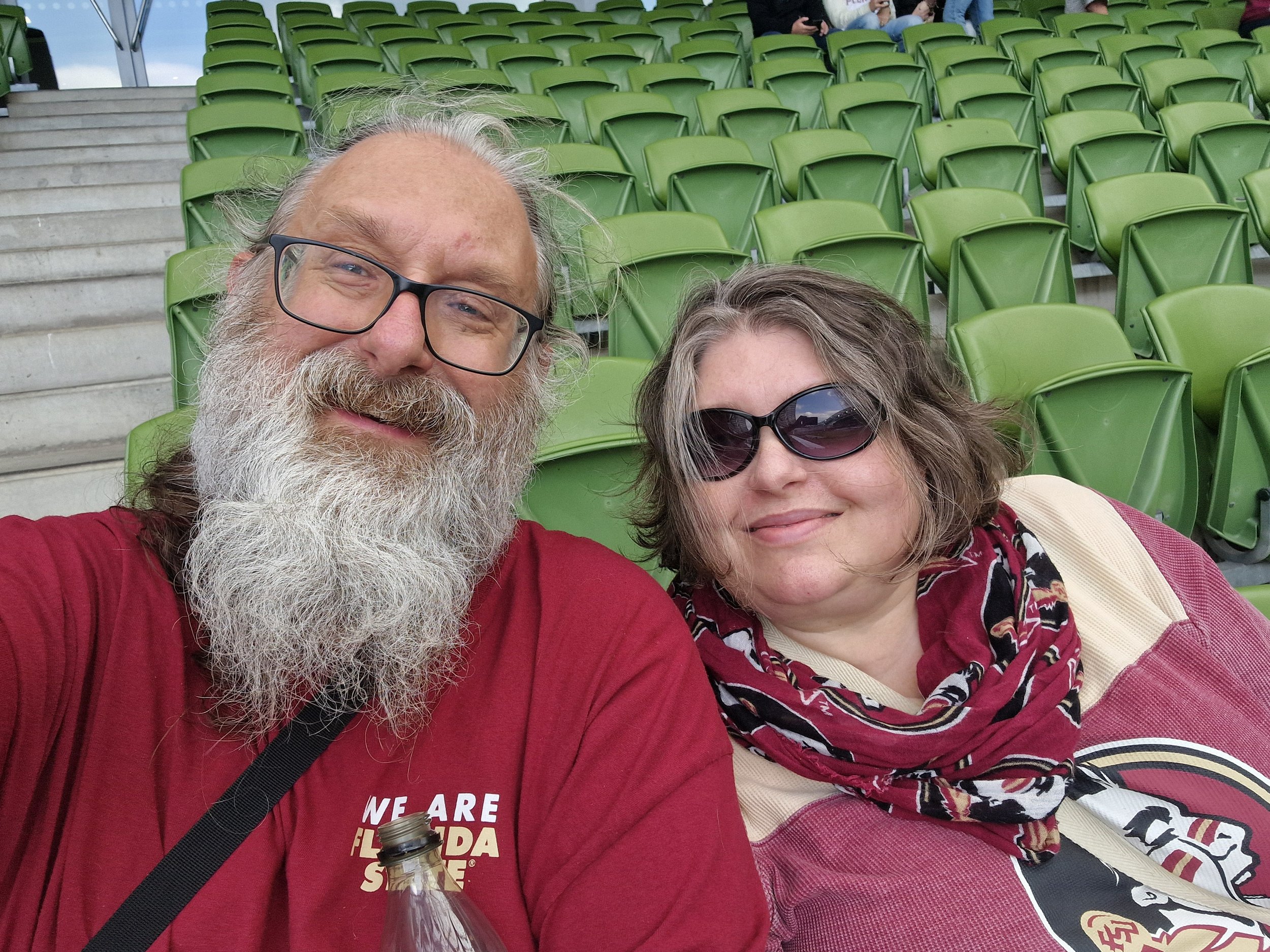A smiling middle-aged man with glasses and a gray beard and a woman with glasses and light brown hair, sitting in a stadium with green seats. The man is wearing a red shirt, and the woman is wearing a patterned scarf and a red top.