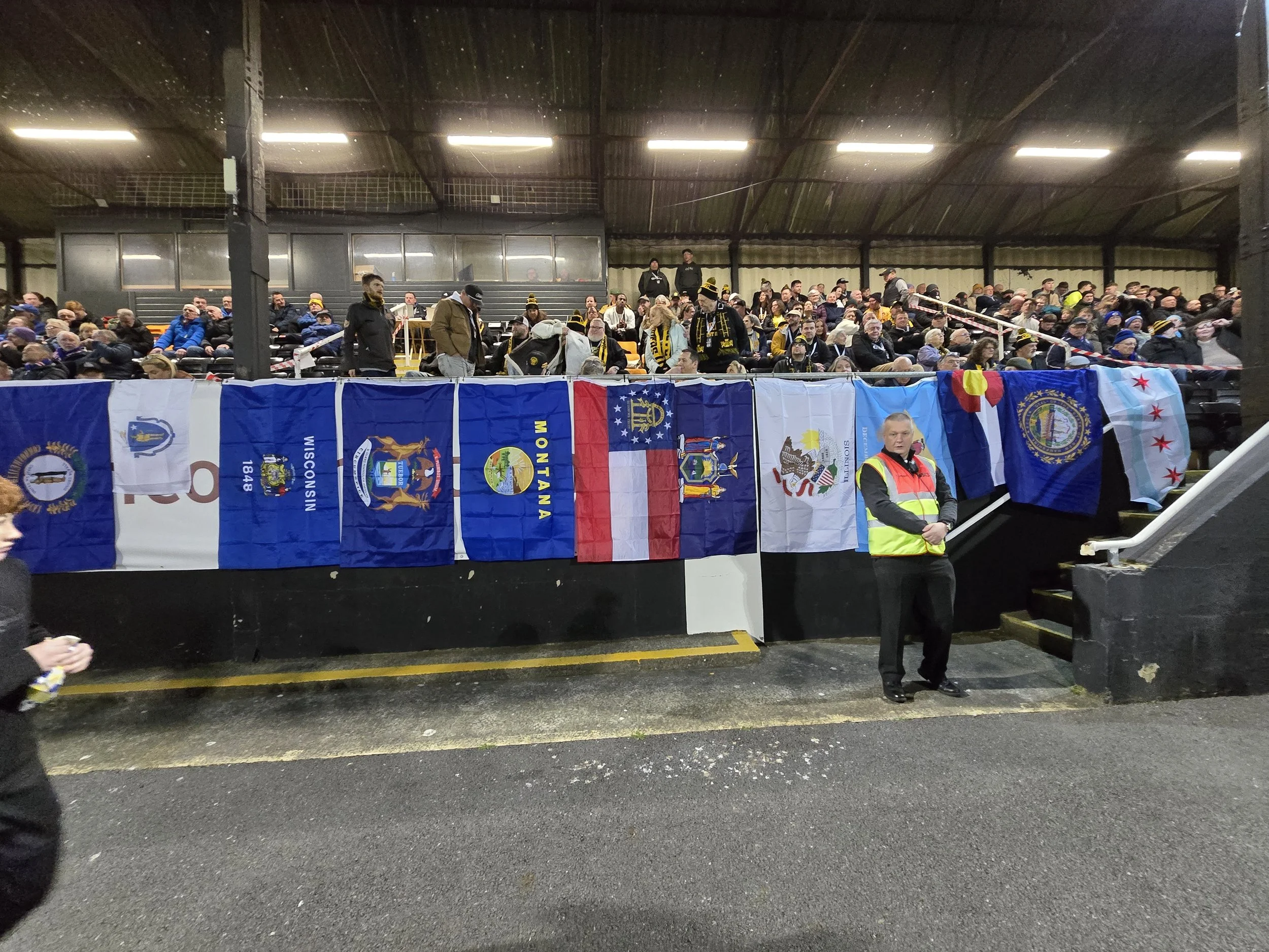 Audience seated in bleachers, many wearing black and yellow scarves and hats, inside a stadium with a dark metal roof, some standing and some seated, with flags hanging from the front of the seating area.