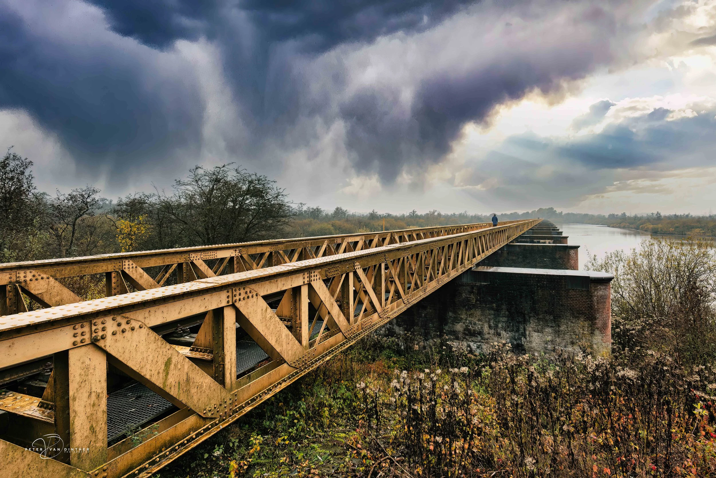 Een oude spoorbrug die over een rivier ligt, onder een bewolkte hemel met dreigende regenwolken. Er is een persoon die op de brug staat, naar de horizon kijkend. Omgeven door herfstbomen met goudgele en groene bladeren.
