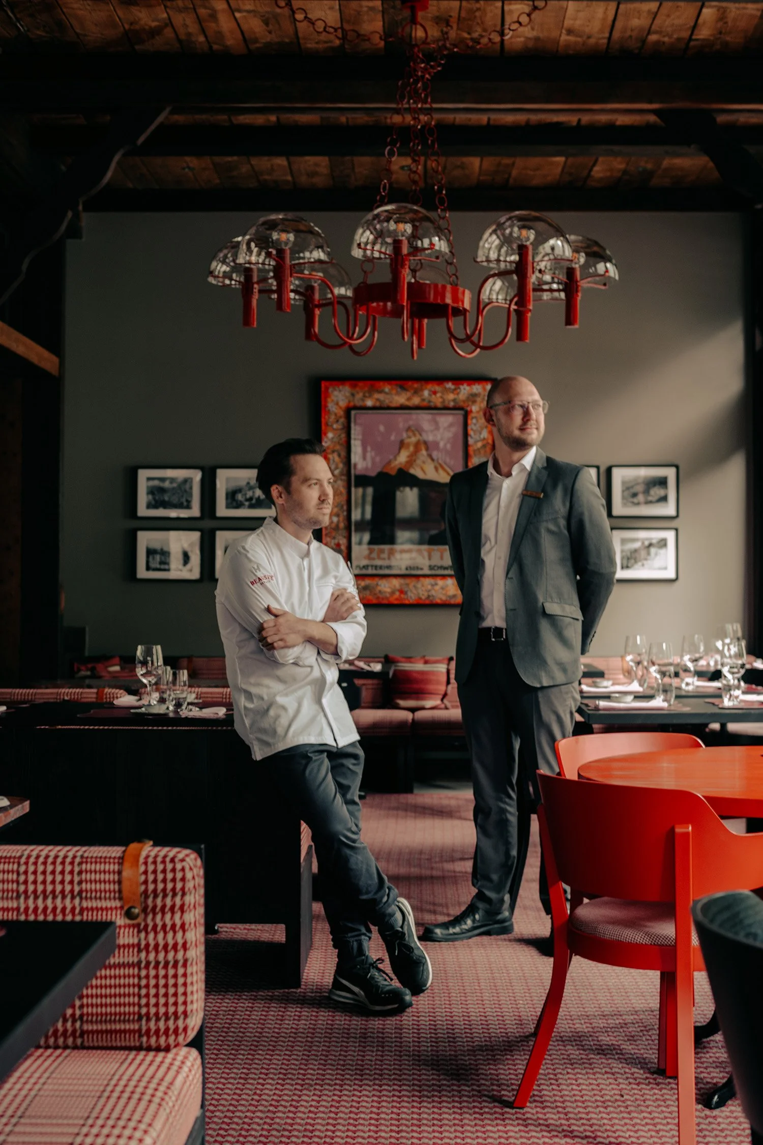 Deux hommes se tiennent debout dans un restaurant : l'un a les bras croisés et l'autre porte un costume. Le restaurant est décoré de tableaux encadrés accrochés aux murs, d'un grand lustre rouge et de chaises rouges.