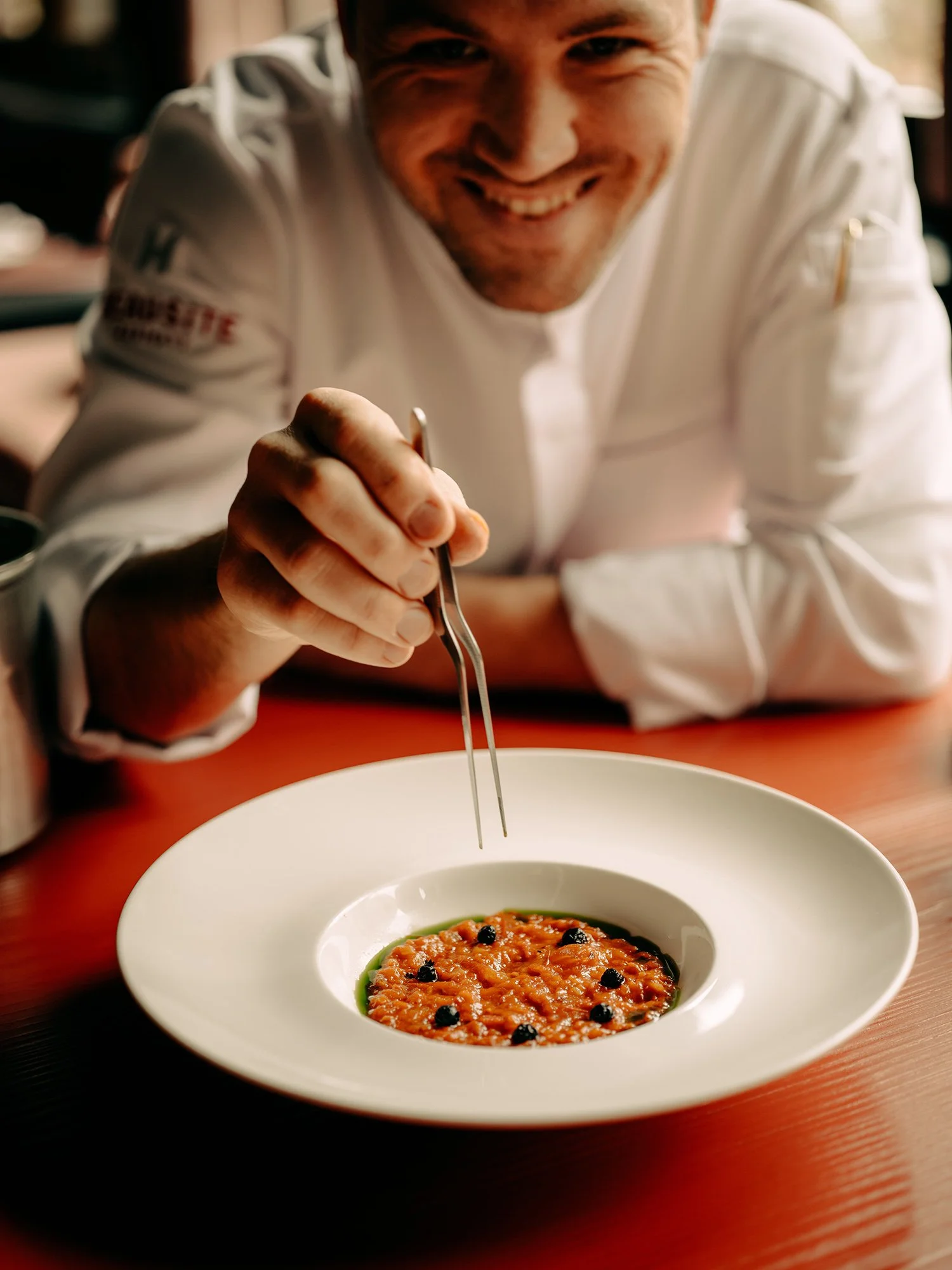 Un chef souriant, vêtu d'un uniforme blanc, utilise une pince pour ajouter une garniture à un bol contenant un plat de couleur orange, peut-être une soupe ou un plat de fruits de mer, posé sur une table.