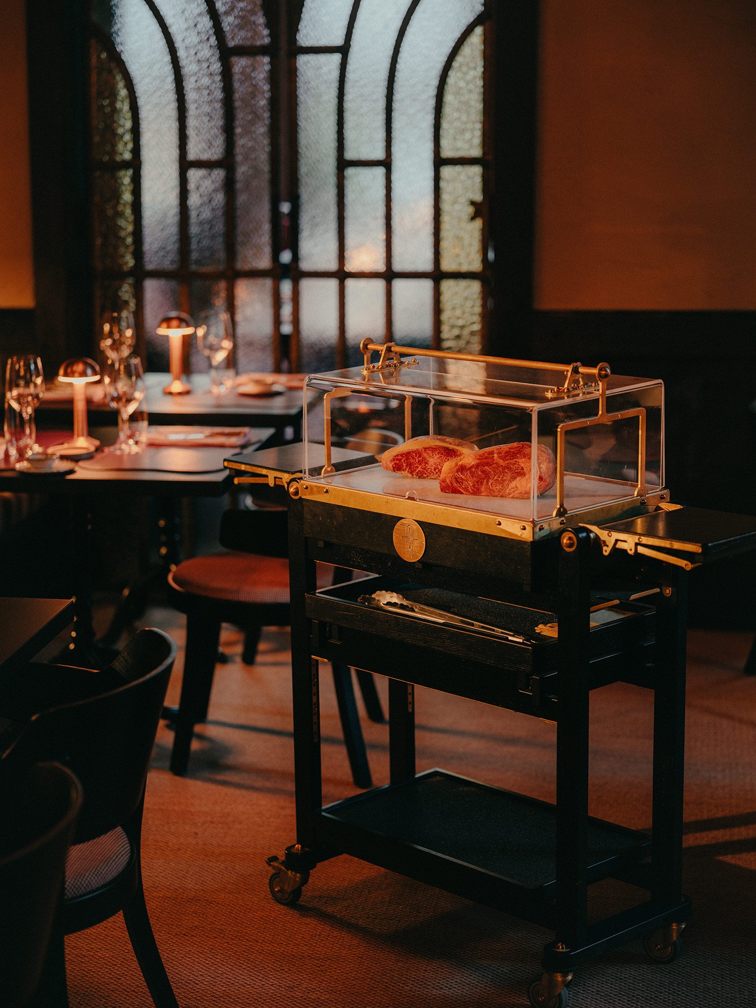 Cadre de restaurant raffiné, avec une vitrine en verre contenant des morceaux de bœuf persillé, posée sur un chariot noir à roulettes, devant un grand vitrail et des tables dressées avec des verres à vin et des bougies.