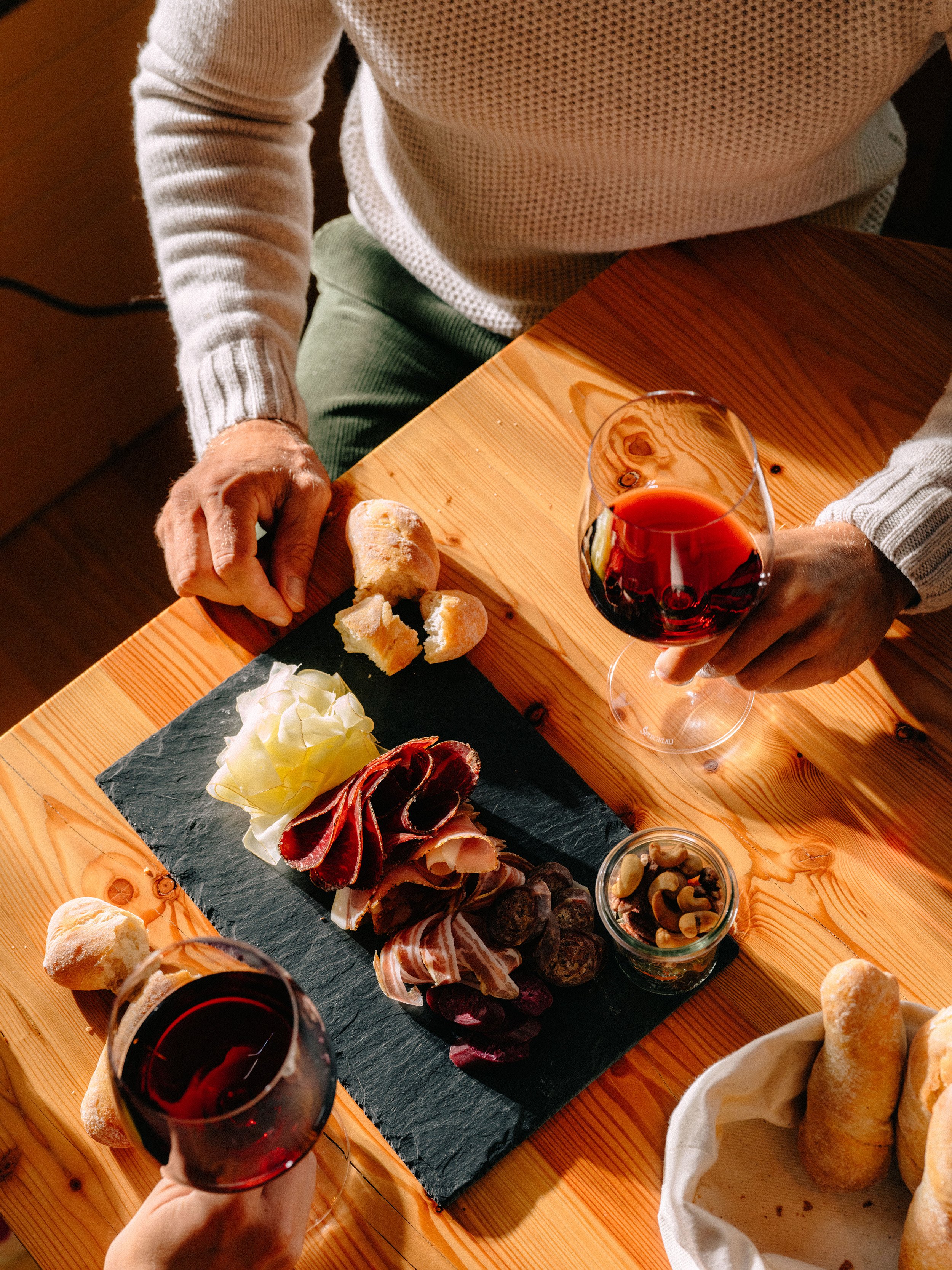 Une personne tenant un verre de vin rouge au-dessus d'une table en bois sur laquelle sont disposés un plateau de charcuterie composé de viandes, de fromages et d'amuse-bouches, ainsi que du pain et un verre de vin rouge.