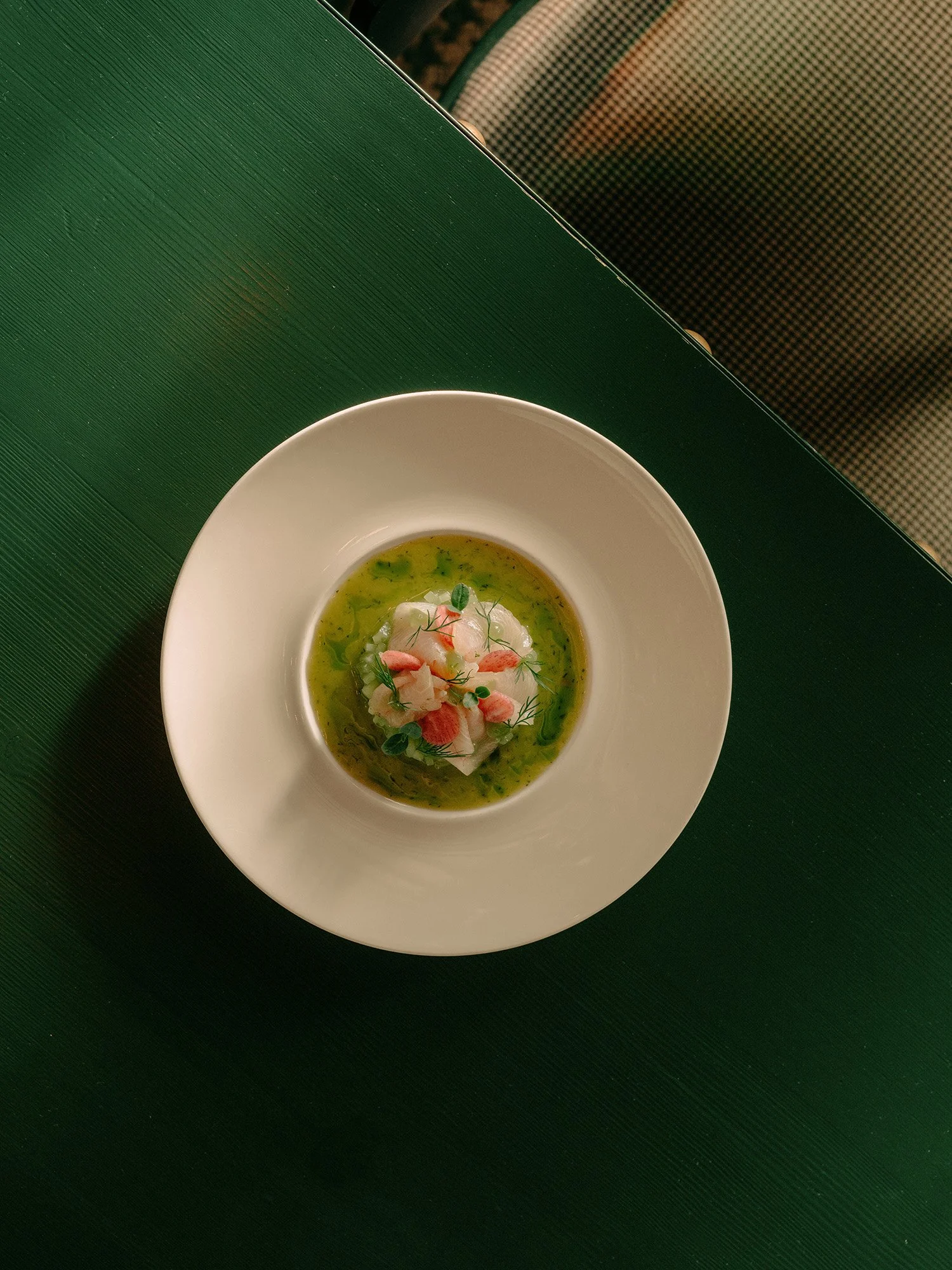 Un bol de soupe verte agrémenté de garnitures, posé sur une table verte texturée, avec une chaise à motifs en arrière-plan.