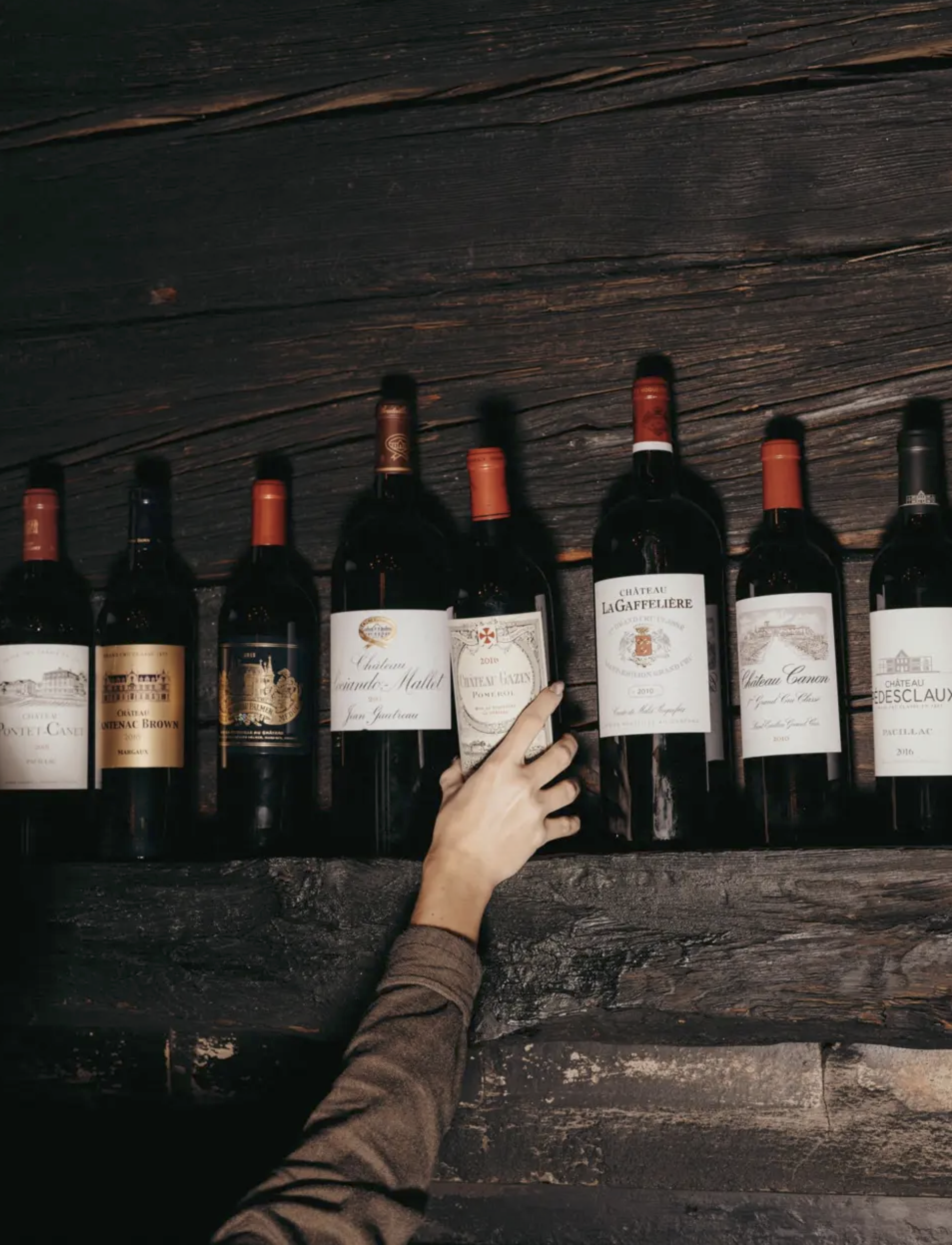 Plusieurs bouteilles de vin rouge alignées sur une surface en bois foncé, avec une main qui saisit l'une d'entre elles.