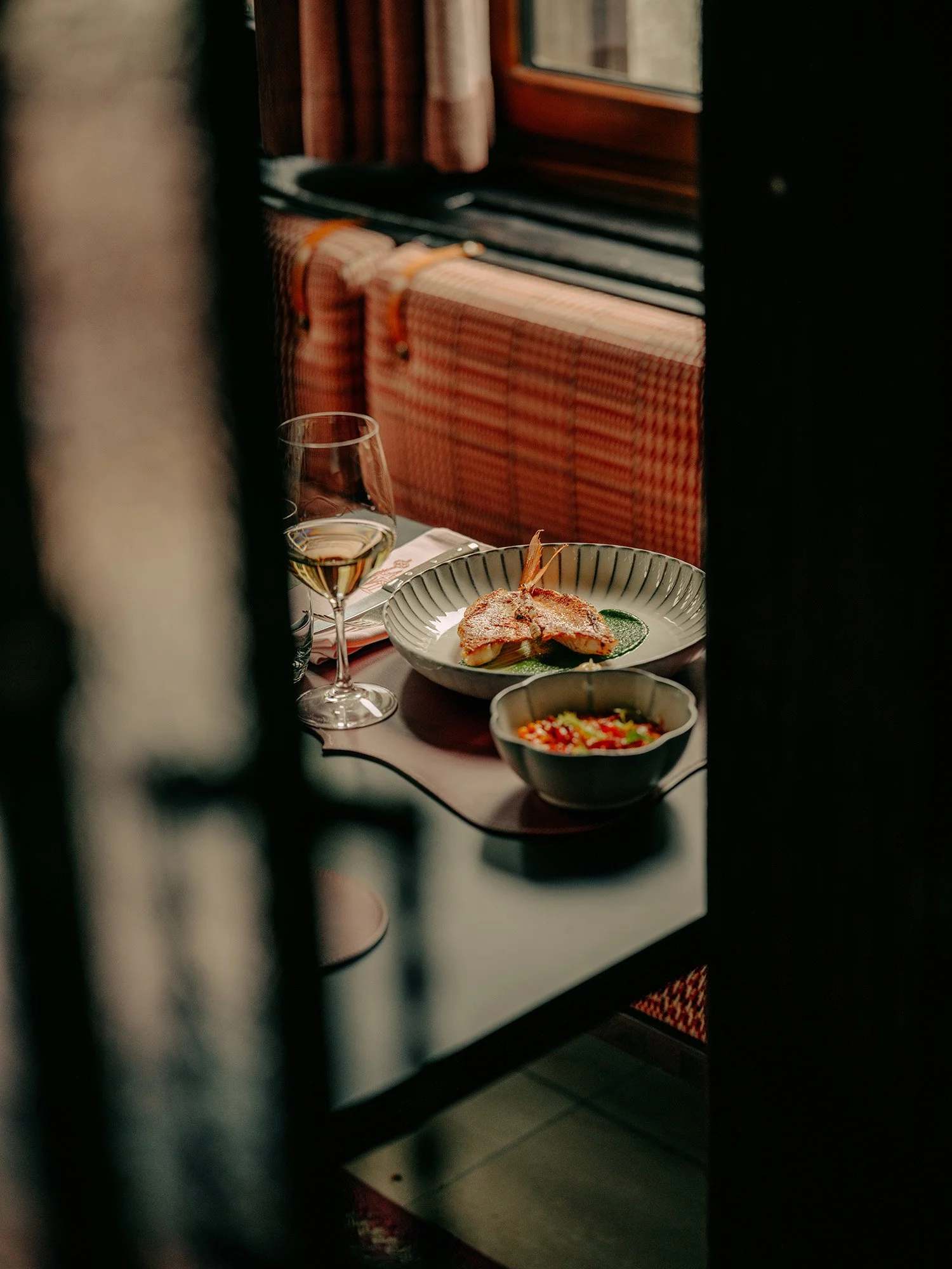 Une table à manger sur laquelle sont posés une assiette de poisson cuit, un verre de vin blanc et un saladier, vue à travers un interstice entre les meubles du restaurant.