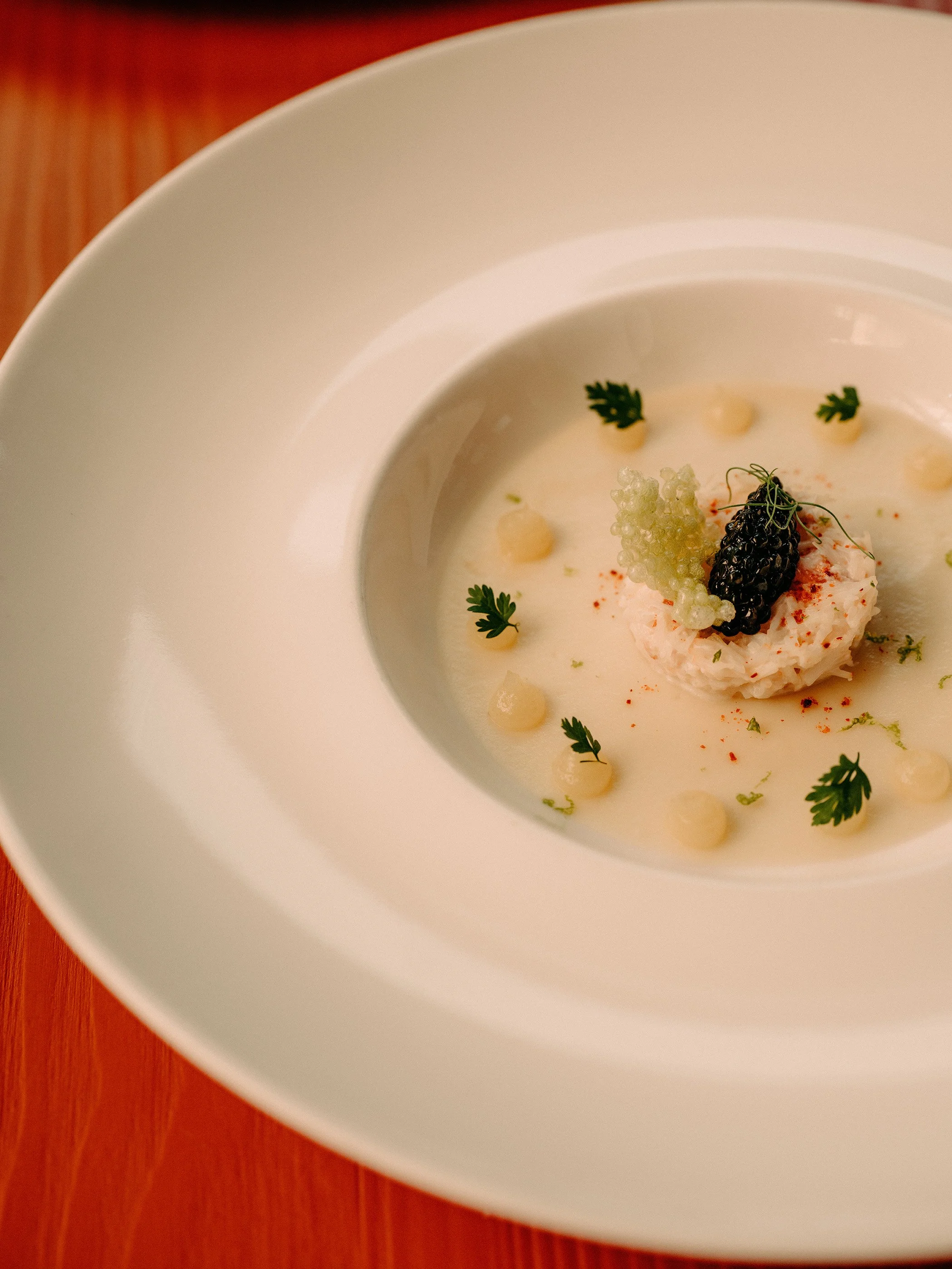 Un bol de soupe onctueuse agrémenté de caviar noir et vert, d'herbes fraîches et de petites cuillerées de sauce, posé sur une table en bois.