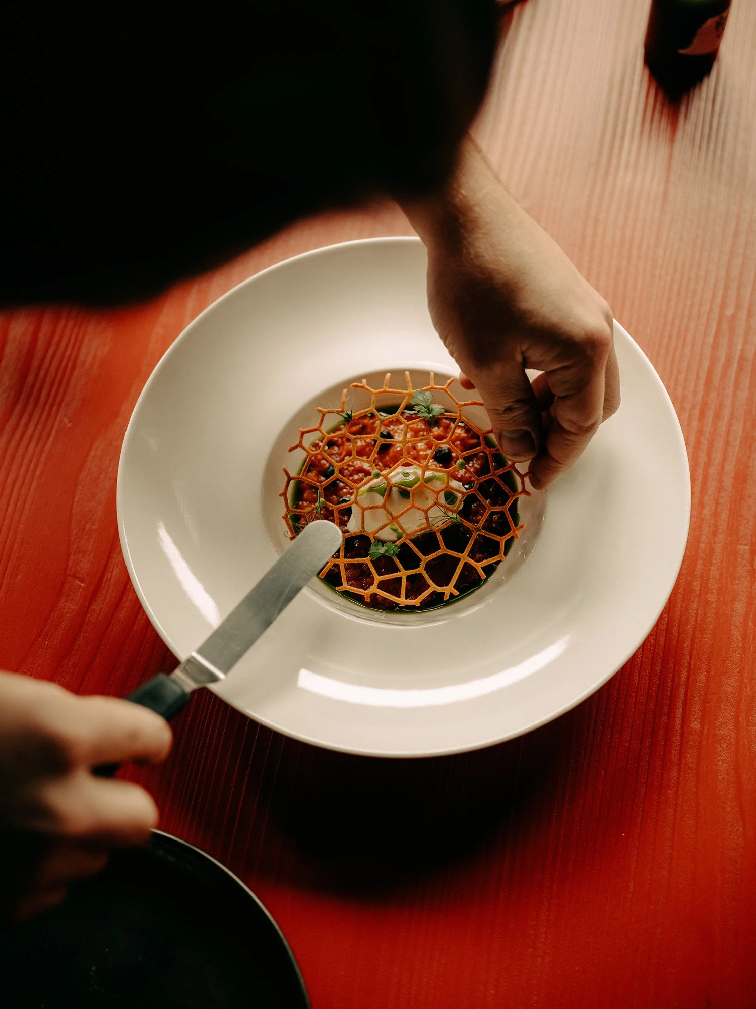 Une main tenant un couteau entame un dessert surmonté d'une structure en nid d'abeille, servi sur une assiette blanche avec une table rouge en arrière-plan.