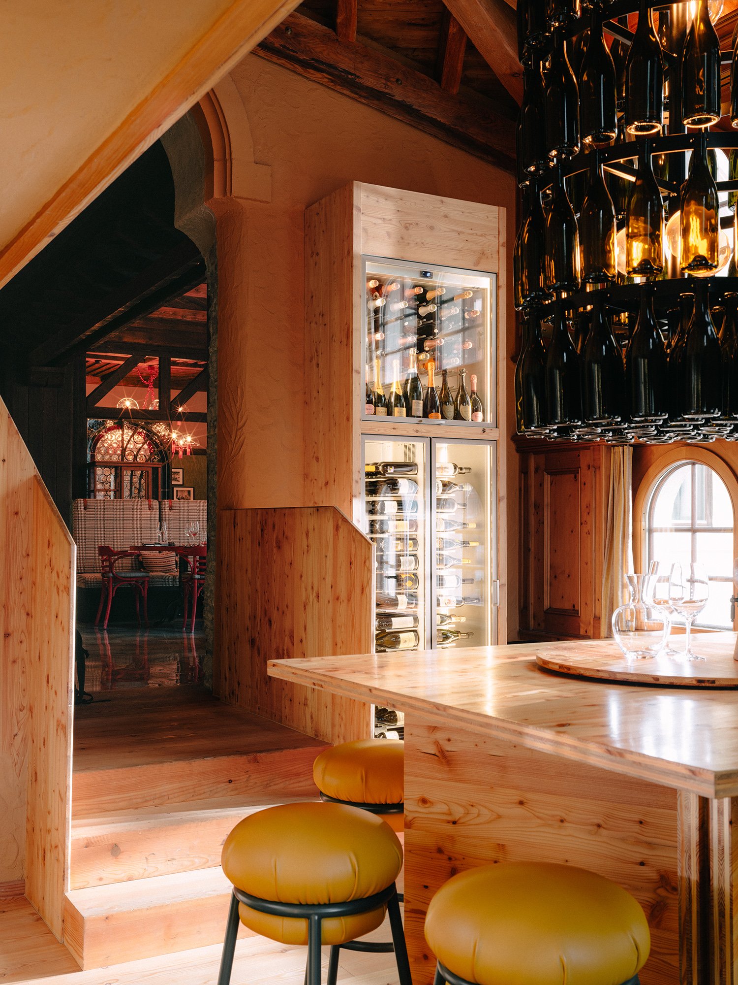 Intérieur chaleureux d'un restaurant en bois, avec une table en bois, des tabourets rembourrés jaunes et une vitrine à vin remplie de bouteilles. On aperçoit à droite un lustre composé de bouteilles de vin suspendues, tandis qu'une fenêtre ornée de rideaux jaunes laisse entrer la lumière naturelle.