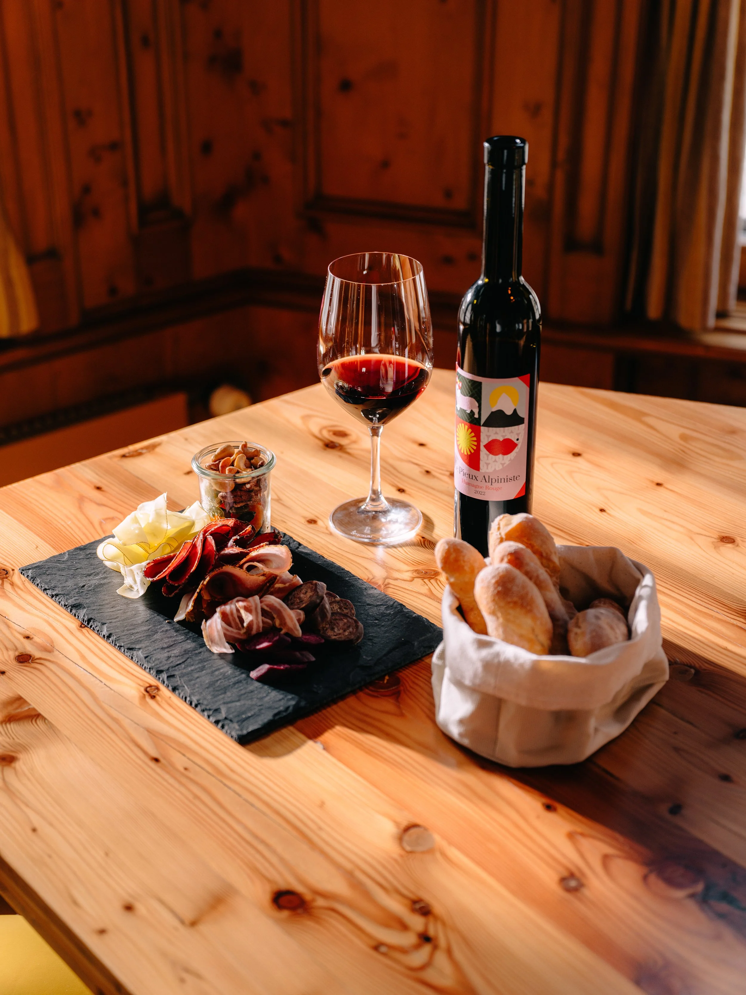 Une table en bois sur laquelle se trouvent un verre de vin rouge, une bouteille de vin à l'étiquette colorée, un petit bocal de noix, un plateau de fromages en tranches et une corbeille de petits pains.