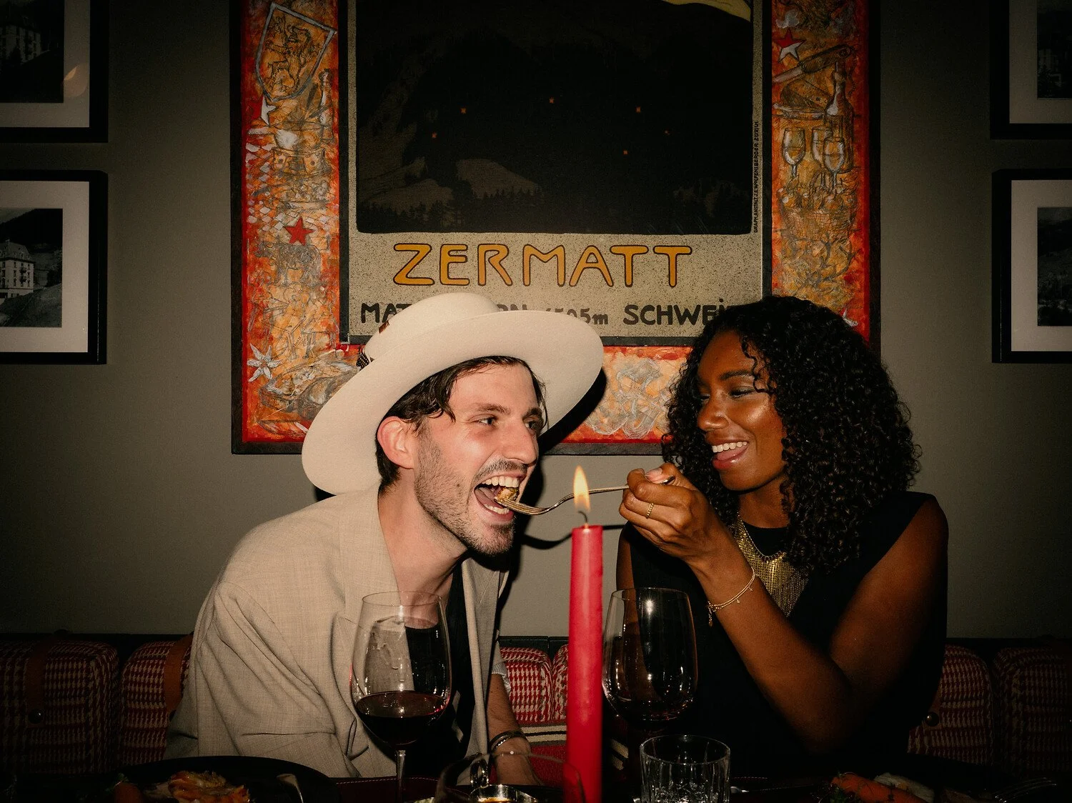 Un homme et une femme font la fête autour d'une table ; l'homme porte un grand chapeau blanc et se sert avec une fourchette, tout en souriant, tandis que la femme lui tend une bouchée à l'aide d'une fourchette ; tous deux ont l'air heureux. Il y a une bougie rouge sur la table et des verres à vin devant eux.