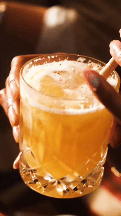 Une personne tenant un verre contenant ce qui semble être un cocktail jaune, avec une paille, dans un décor faiblement éclairé.