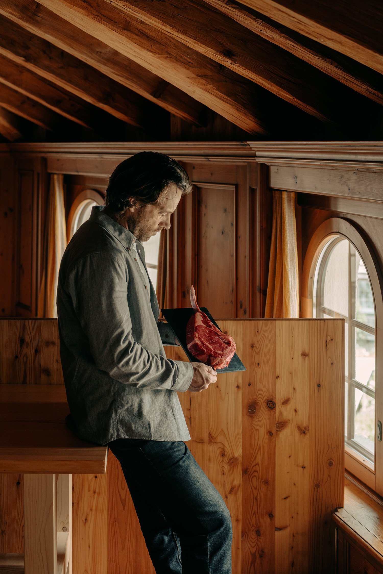 Un homme aux cheveux et à la barbe foncés, vêtu d'une chemise grise et d'un jean, debout à l'intérieur d'une cabane en bois baignée par la lumière naturelle qui pénètre par la fenêtre, tenant un morceau de viande crue.