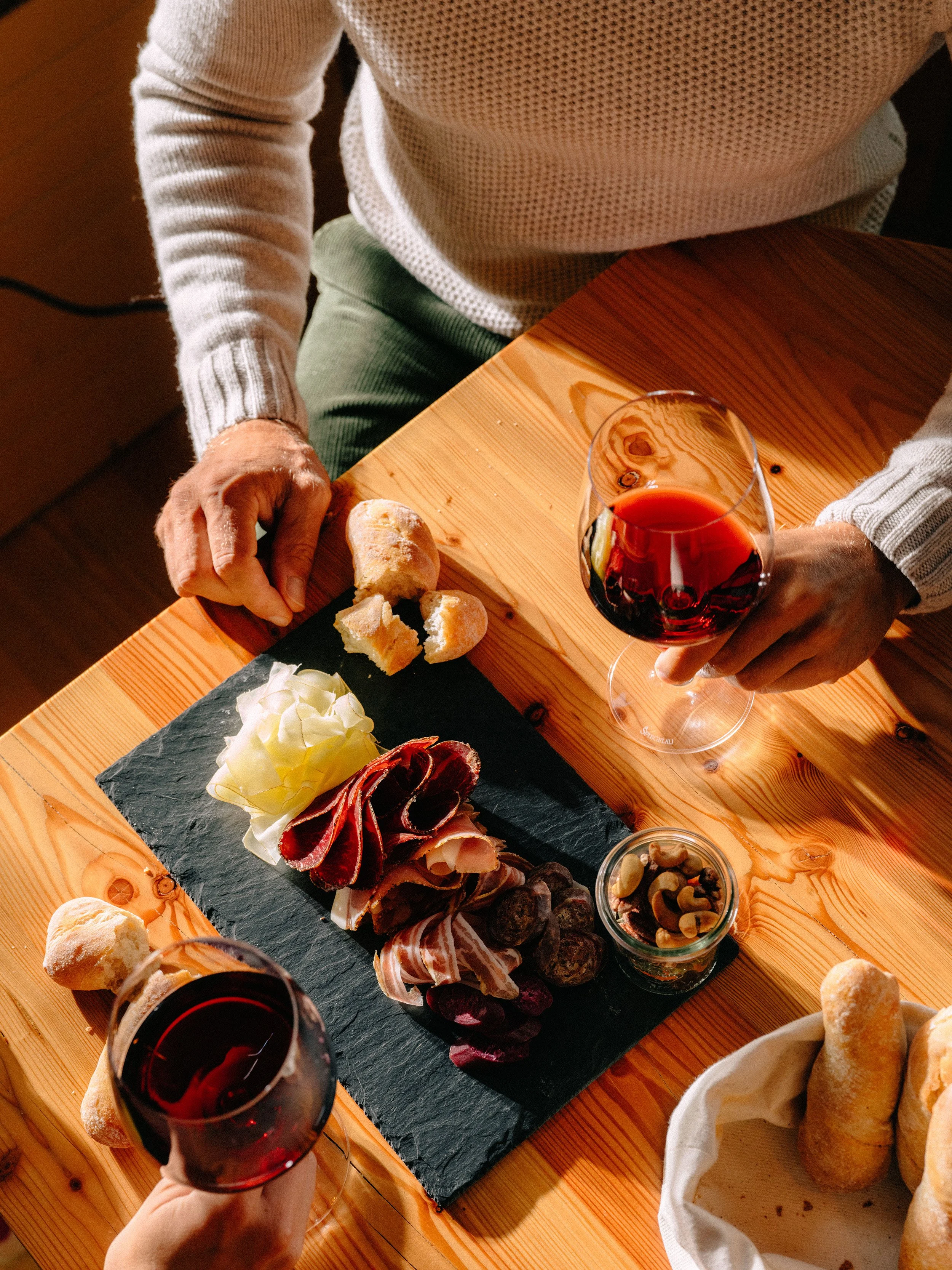 Une personne tenant un verre de vin rouge, assise à une table en bois devant un plateau de charcuterie composé de charcuterie, de fromage, de crackers et de fruits secs.