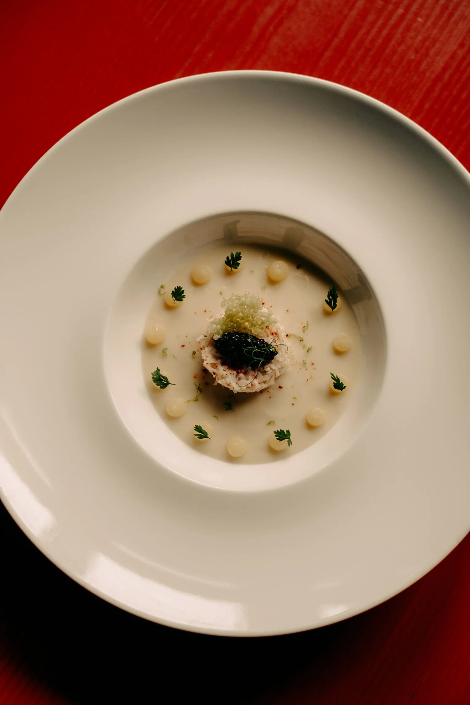 Un bol blanc contenant un plat crémeux agrémenté de petites herbes, d'une sauce noire et de cuillerées de sauce, posé sur un plateau en bois rouge.