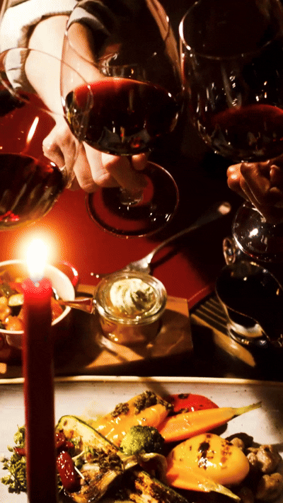 Plusieurs verres de vin rouge en train d'être versés ou tenus à la main, avec une bougie allumée et une assiette de légumes rôtis posés sur une table rouge foncé.