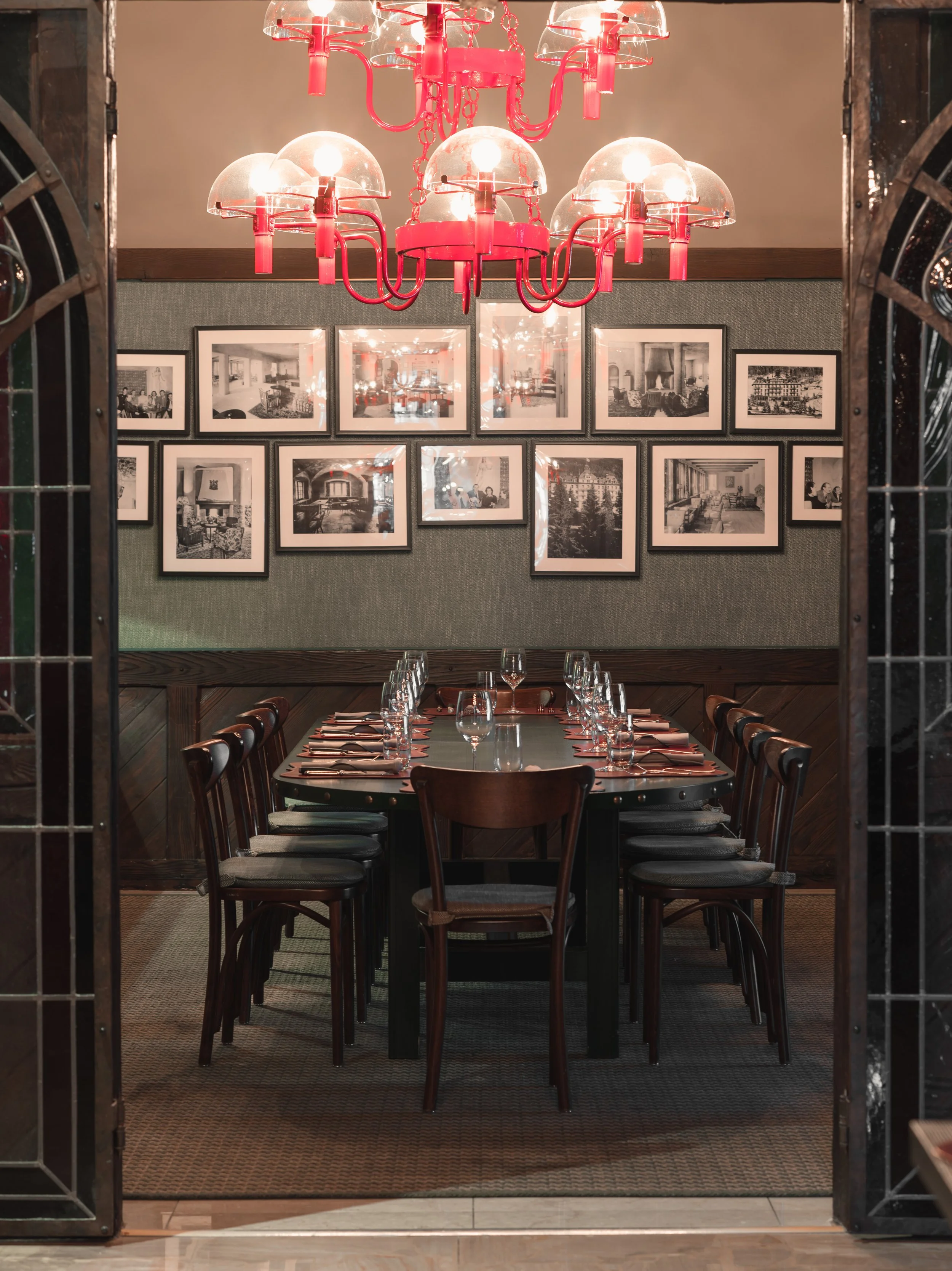 Une salle à manger avec une longue table dressée, sur laquelle sont disposés des verres à vin et des serviettes. Un grand lustre rose, composé d'ampoules apparentes, est suspendu au plafond. Le mur du fond est orné de photographies en noir et blanc encadrées représentant des intérieurs.