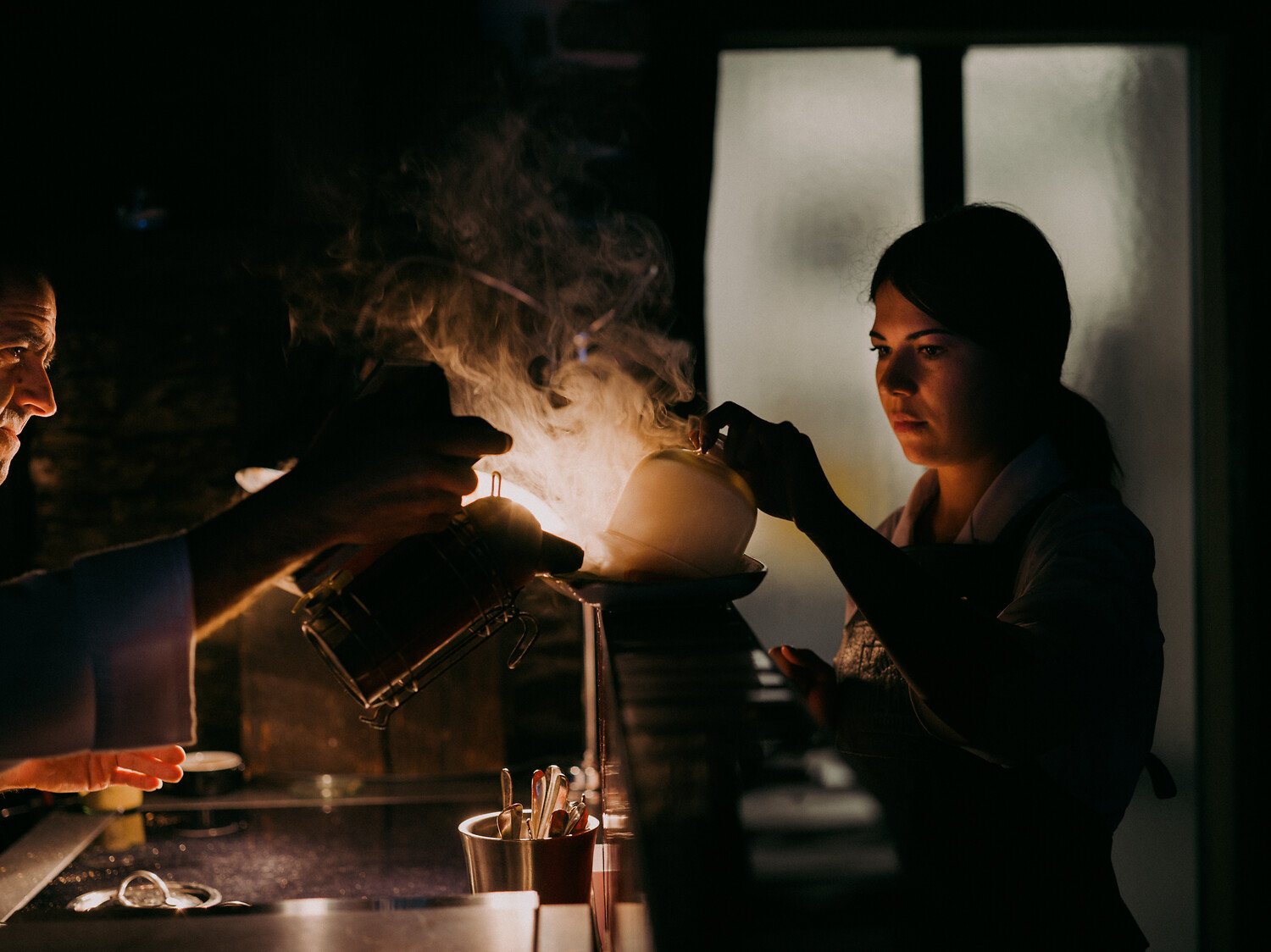 Deux femmes travaillant dans une cuisine faiblement éclairée, penchées au-dessus d'une casserole fumante et d'une bouilloire posées sur la cuisinière, avec une fenêtre en arrière-plan.
