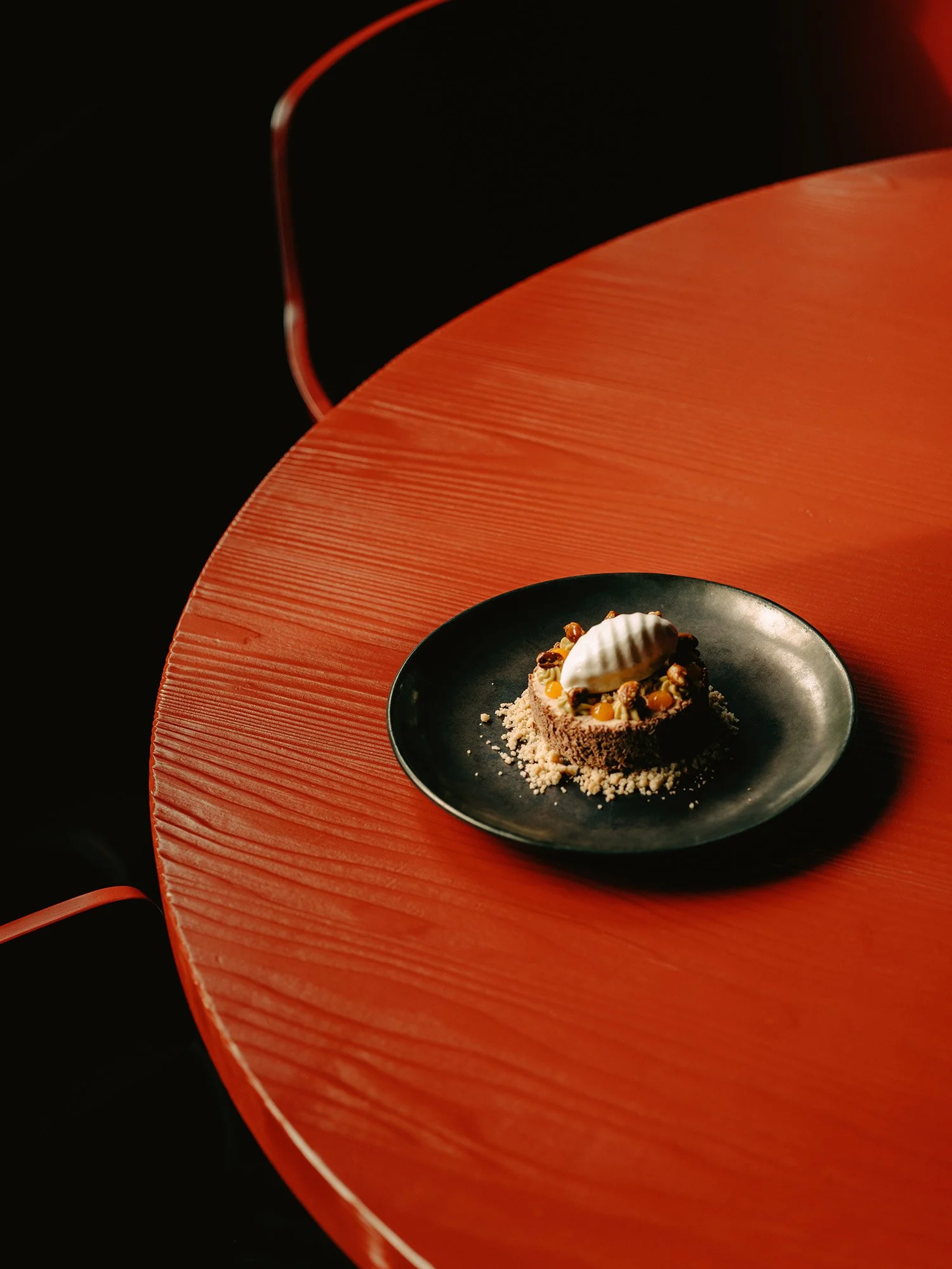 Une assiette noire sur laquelle est disposé un dessert raffiné, agrémenté de noix, de crème et d'écorce d'orange, posée sur une table rouge texturée.