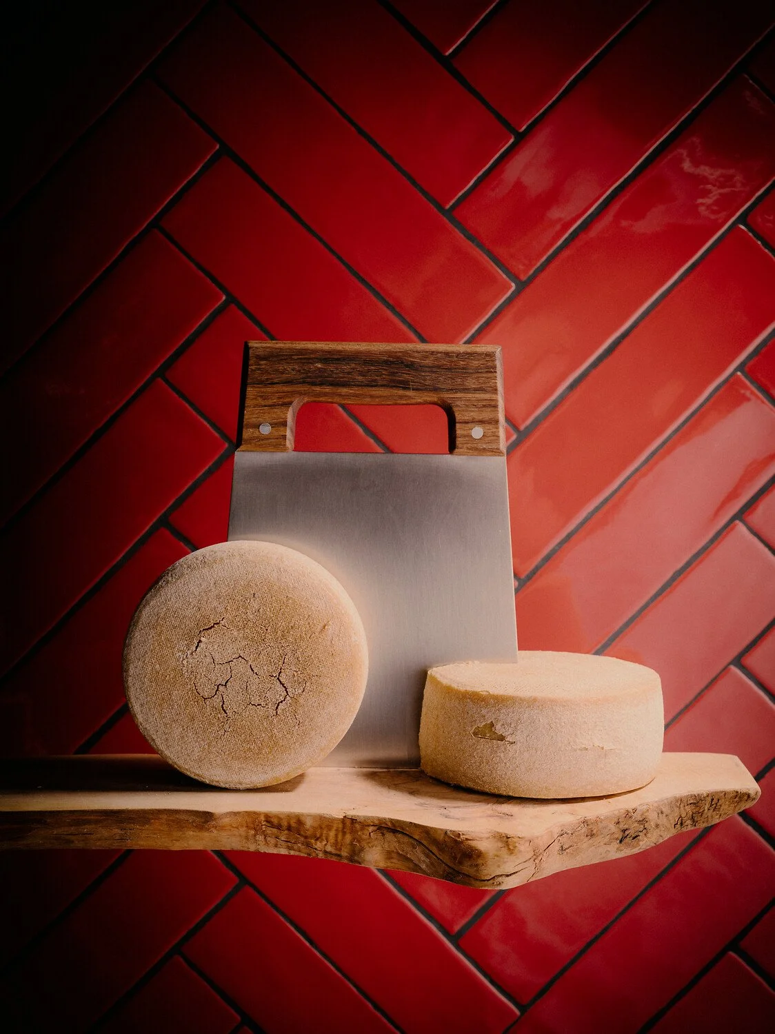 Deux meules de fromage, l'une à la croûte fissurée et l'autre percée d'un trou, posées sur une étagère en bois adossée à un mur de briques rouges, avec un couteau à fromage derrière elles.