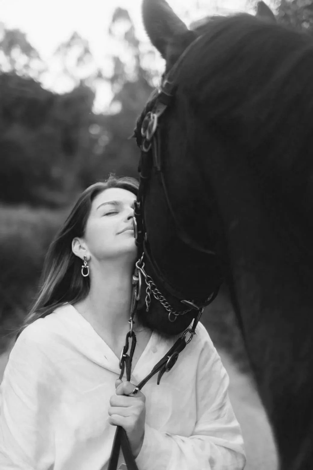 A woman with closed eyes and long hair, wearing earrings and a light blouse, stands close to a large black horse outdoors, gently holding its reins and leaning her face against the horse's nose.