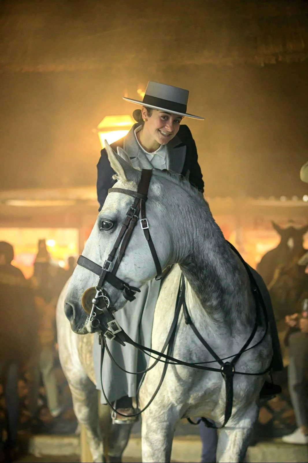 A woman riding a gray horse, wearing a wide-brimmed hat and formal riding attire, indoors with a warm lighting and blurred crowd in the background.