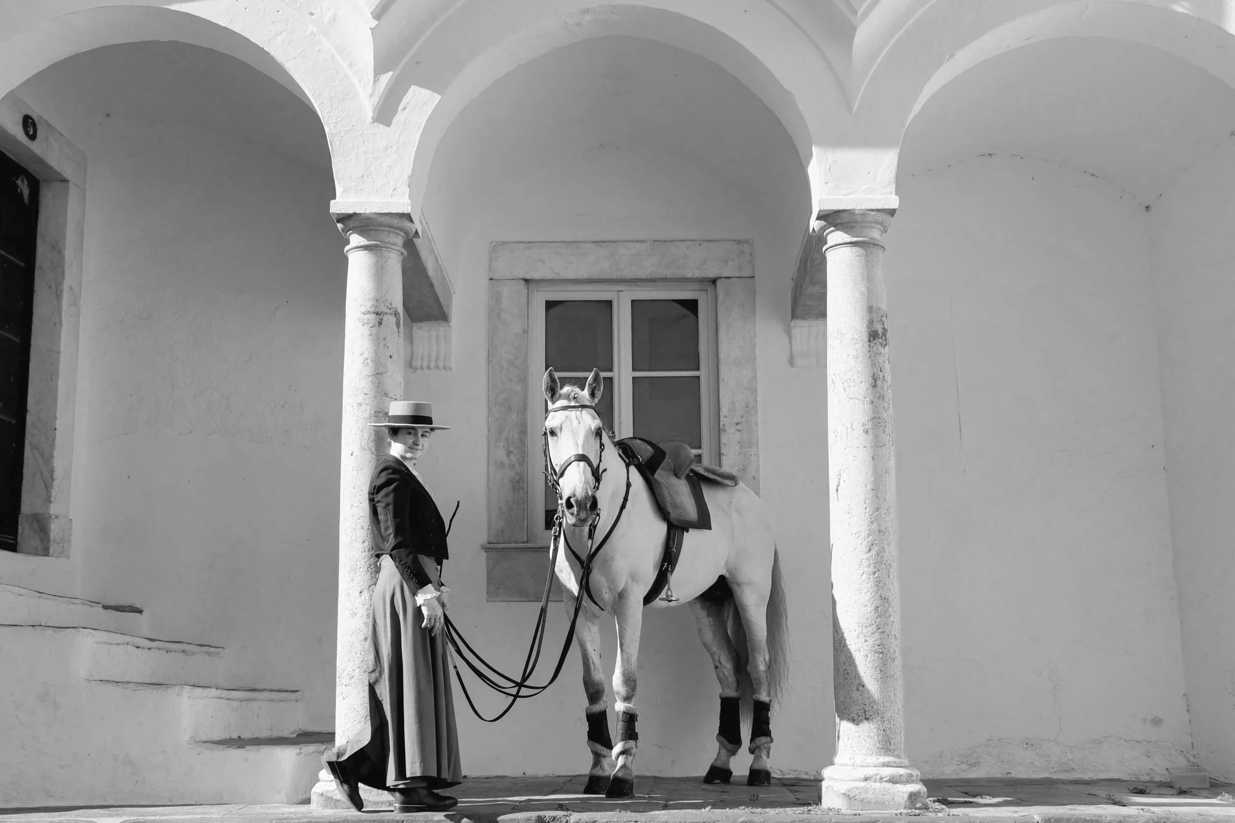 A person in formal attire with a wide-brimmed hat stands next to a white horse with saddles and bridles, in front of an arched doorway supported by columns on either side.