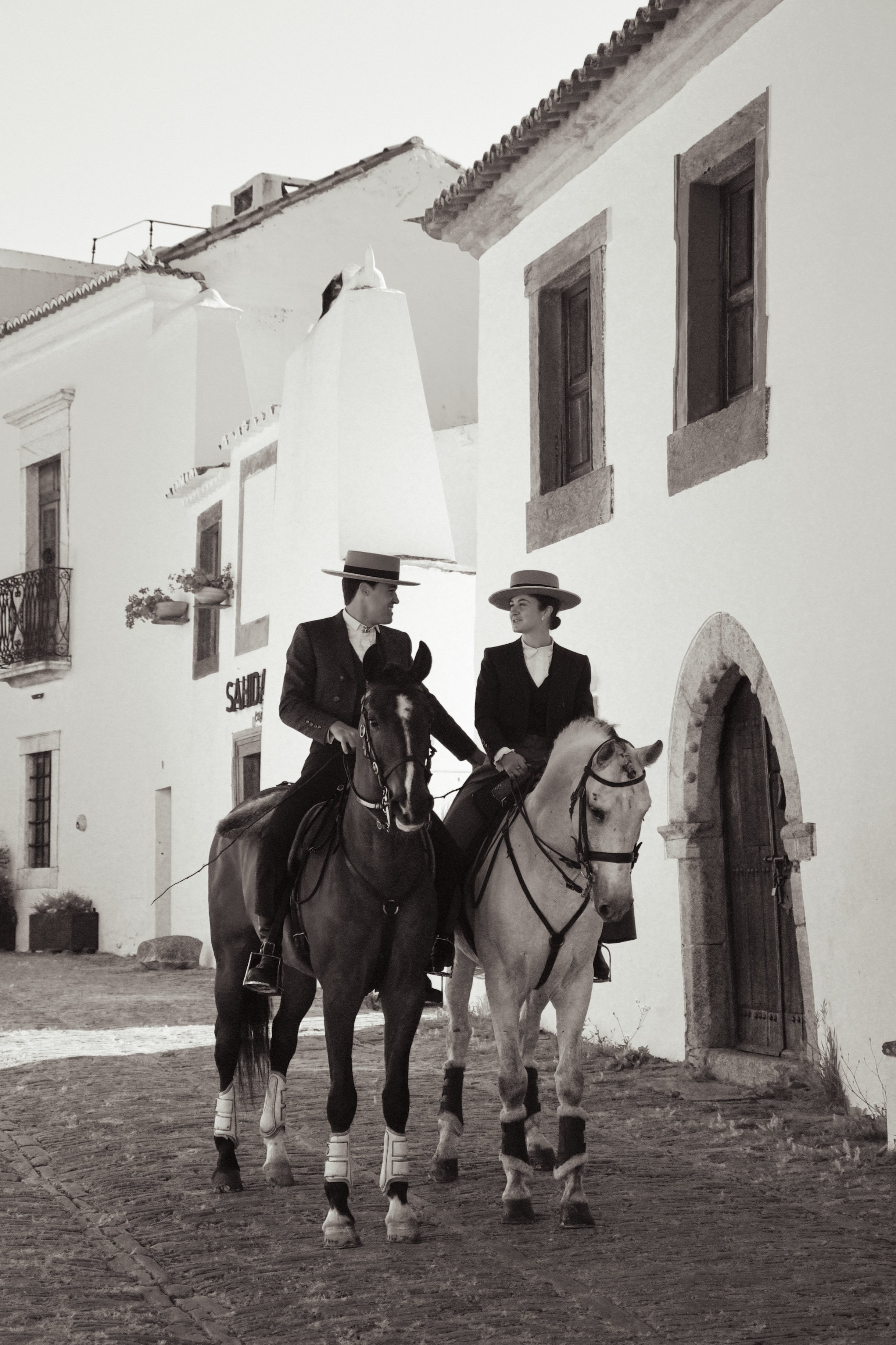 Two women riding horses in front of a white building with windows and shutters, during daytime.