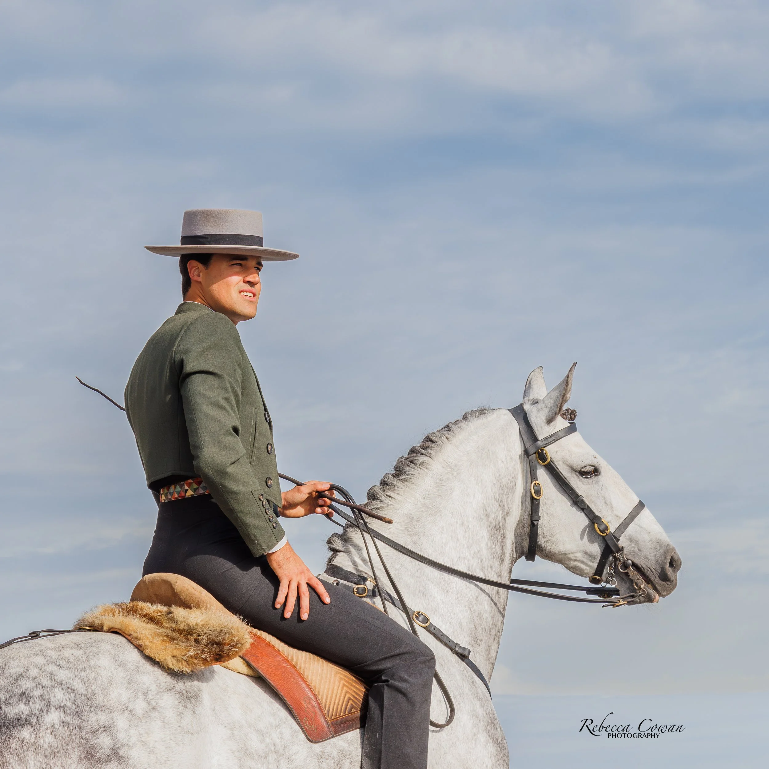 A person riding a white horse with a dark mane, holding the reins, against a cloudy sky. The person is wearing a gray wide-brimmed hat, a gray coat, and black pants. The horse has a brown saddle with a fleece pad underneath. The person appears to be outdoors, perhaps during a horseback riding activity.