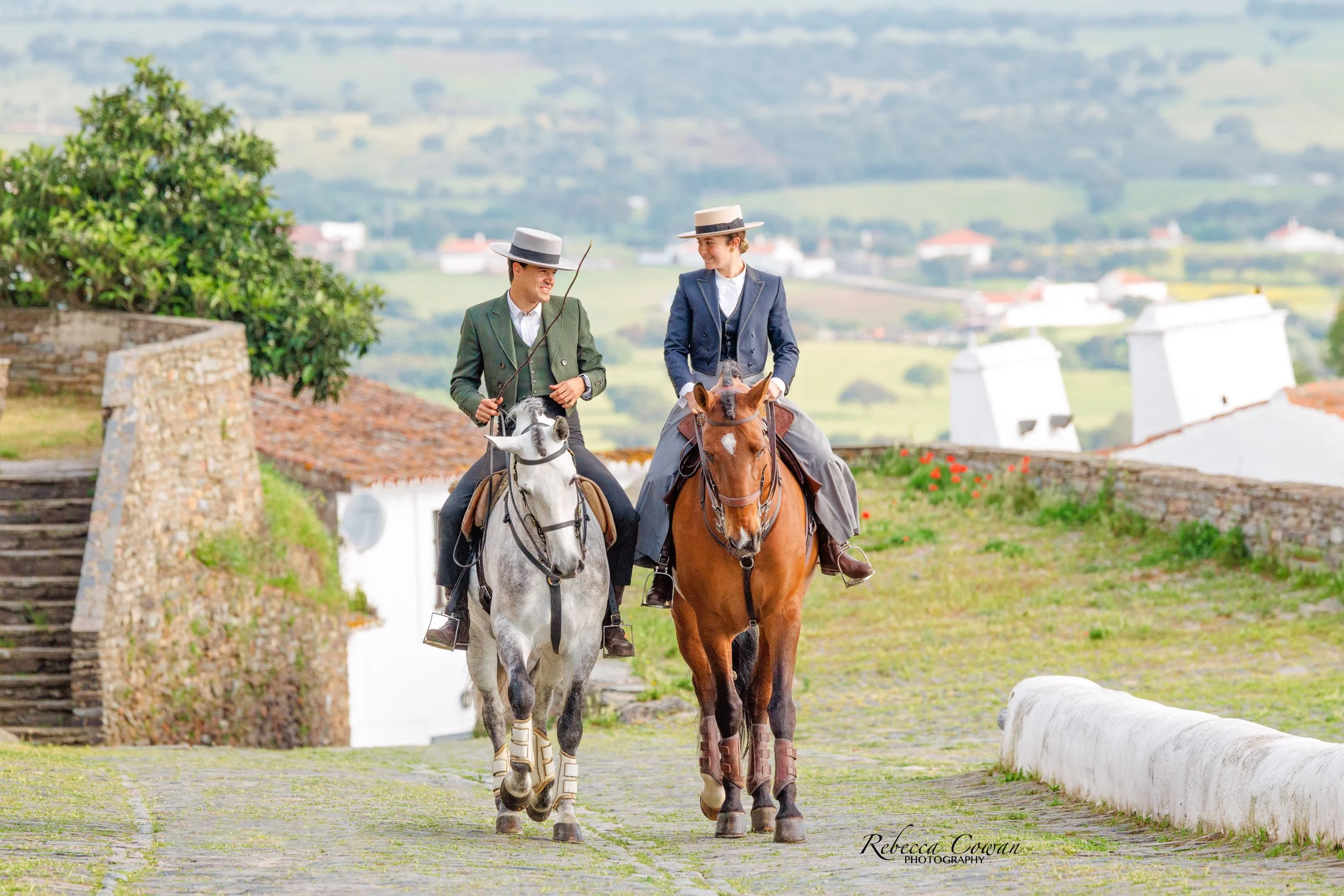 Two women riding horses down a cobblestone path in a scenic countryside with rolling hills and houses in the background, both wearing vintage-style riding jackets and wide-brimmed hats.