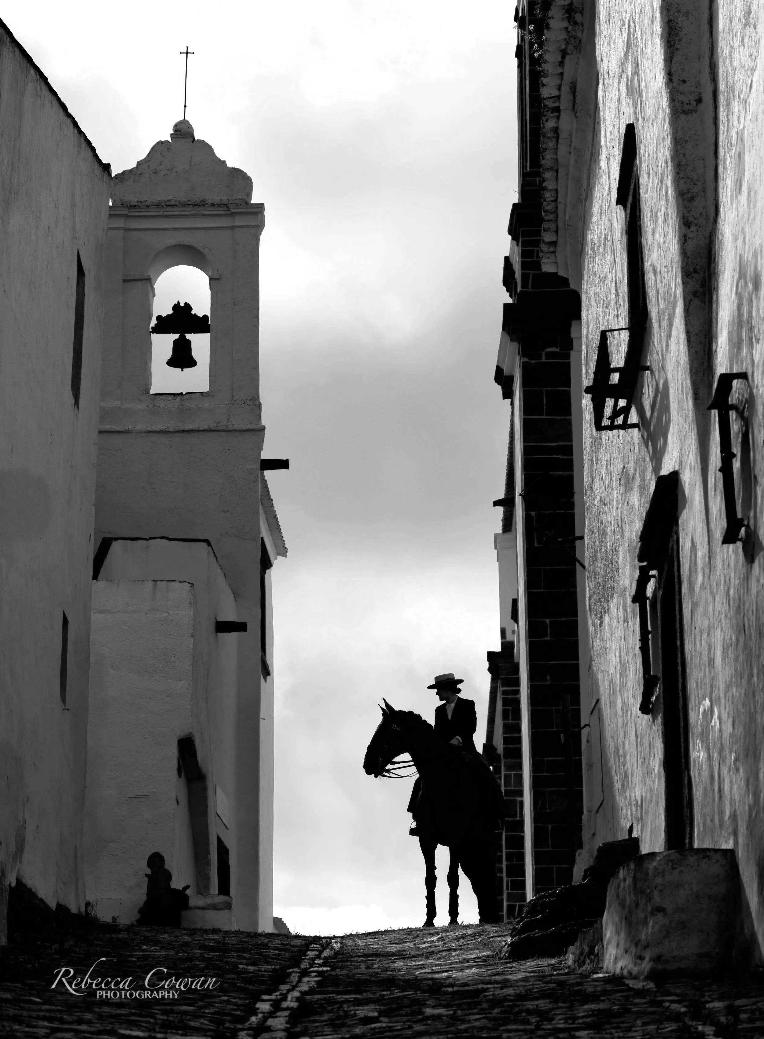 A black and white photo of a person riding a horse along a cobblestone street, with the silhouette of a church tower and a small bell in the background. The scene appears historic and peaceful, with shadows cast by the setting or rising sun.