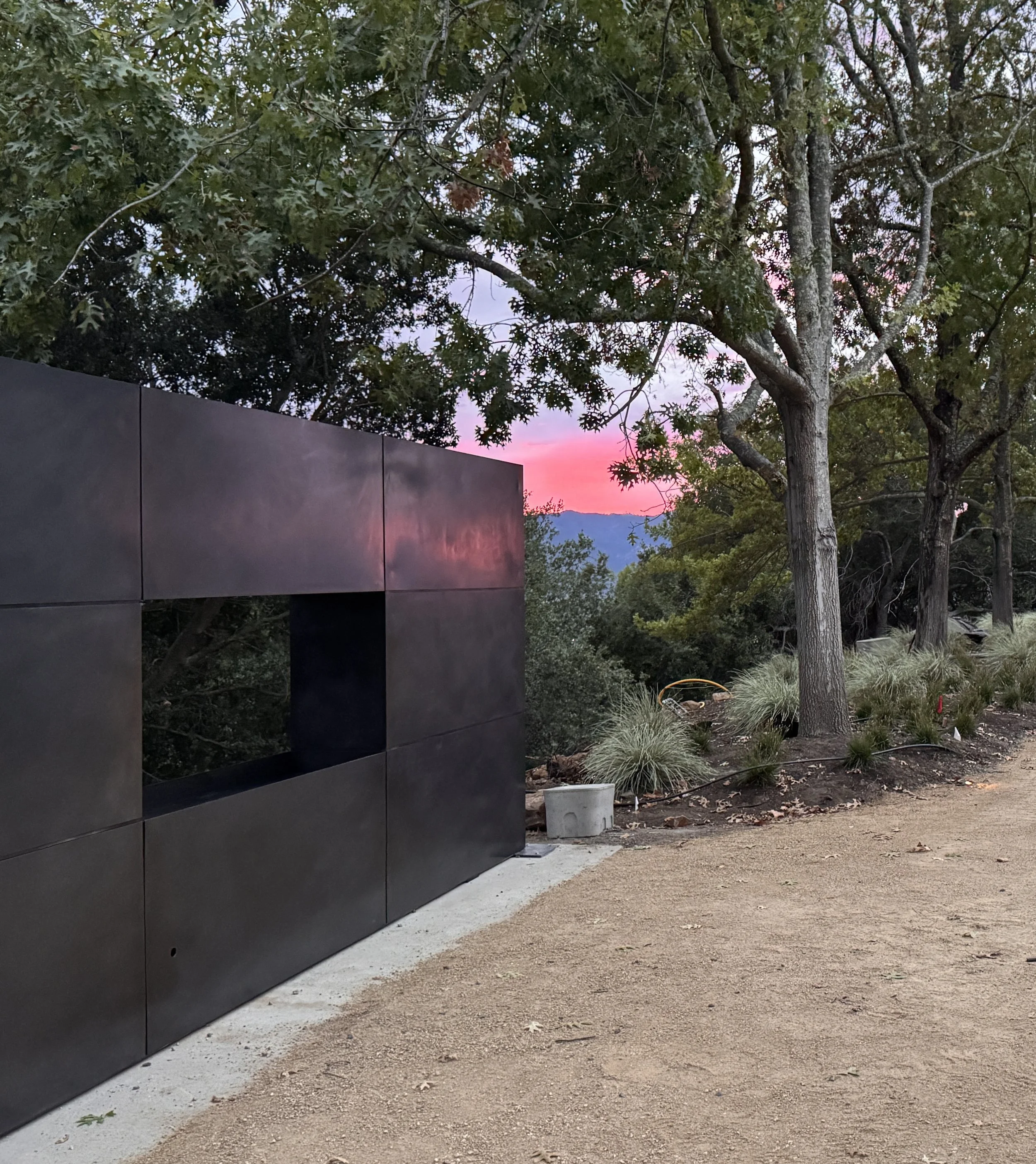 A modern architectural blackened stainless steel fireplace, trees with green leaves and a pink sunset sky in the background, a dirt path with fallen leaves, and some shrubs and grass on the ground.