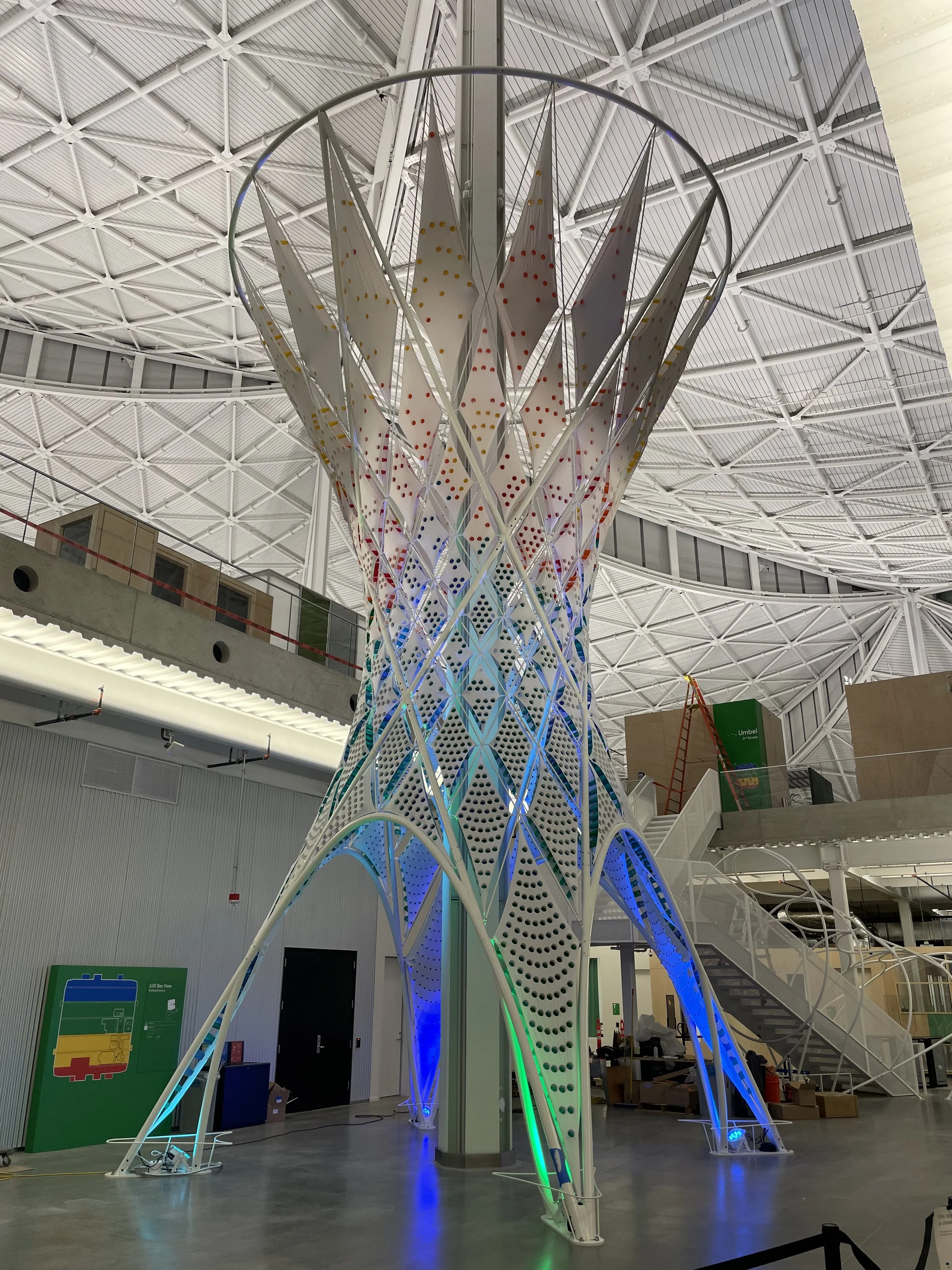 An art installation featuring a tall, white lattice structure with colorful spots, inside a spacious modern building with a high ceiling and metal framework, illuminated with blue, green, and white lighting.