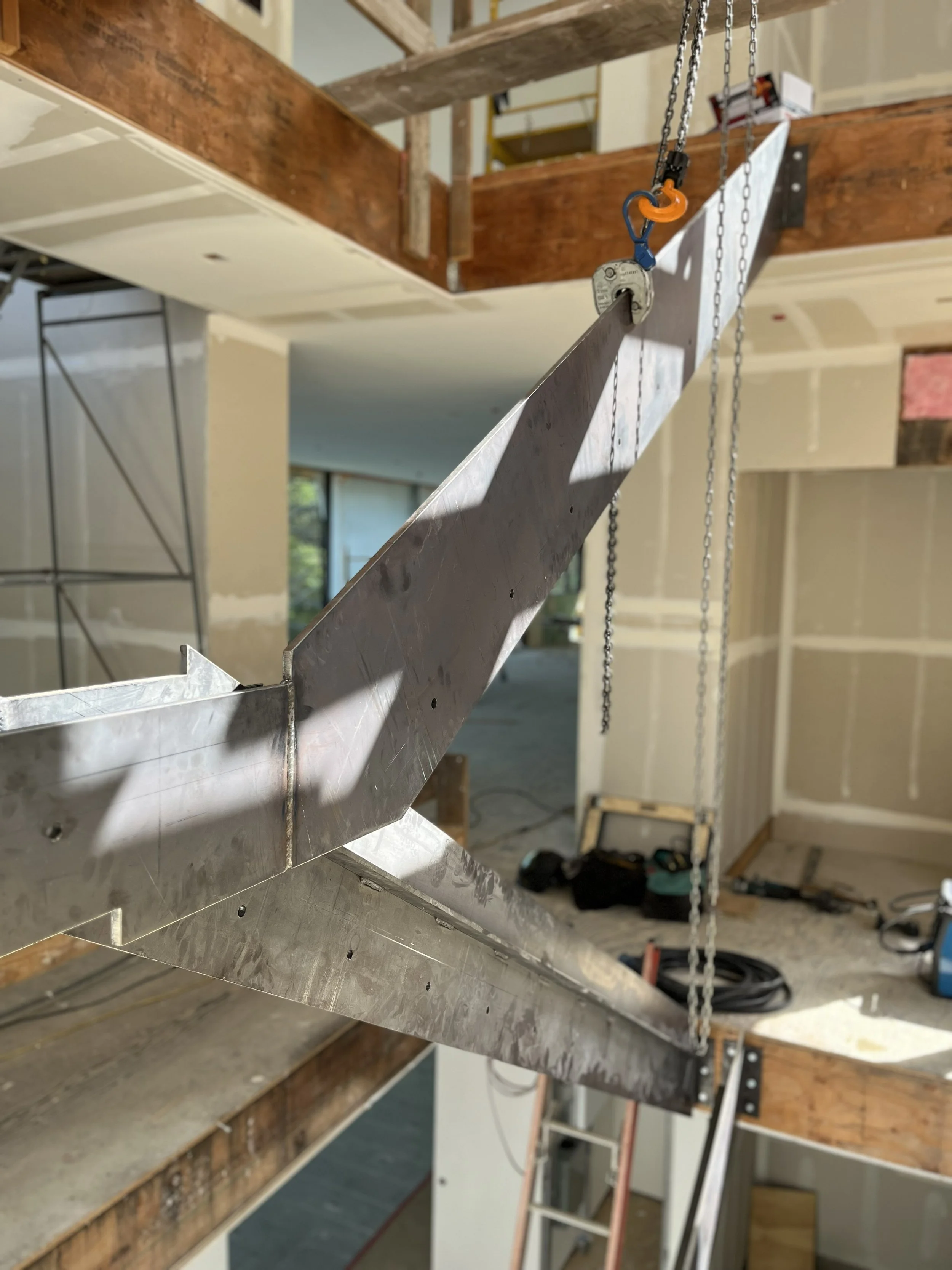 Close-up of a metal beam with a chain hoist attached, in an unfinished interior space with wooden framing and construction tools in the background.