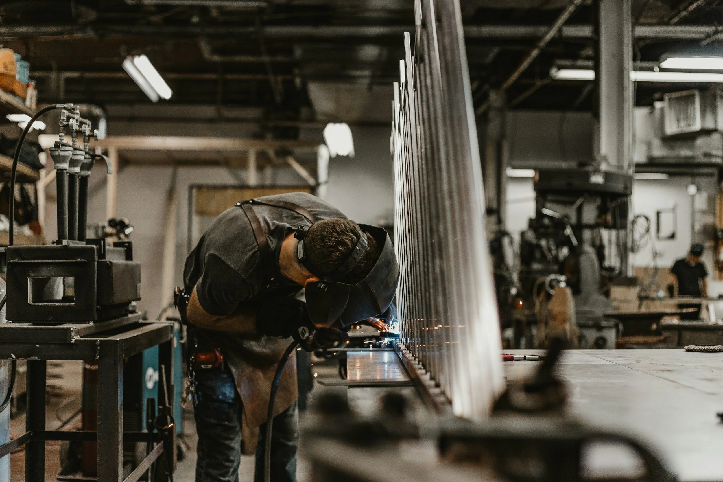A worker welding metal in a workshop with various tools and equipment around.