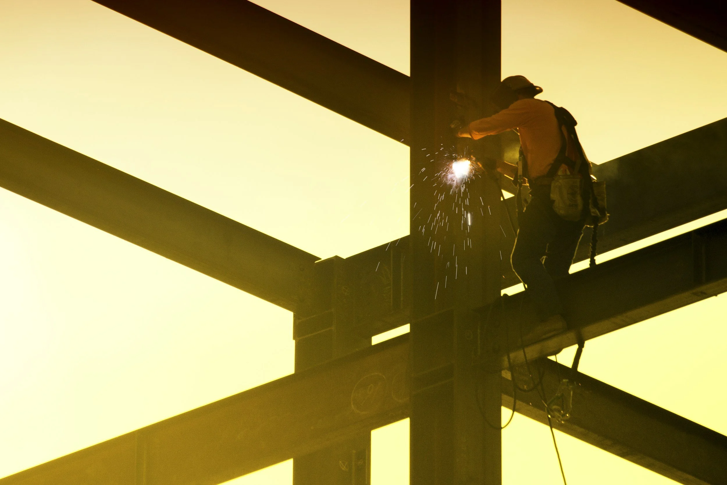 A construction worker welding on a steel structure during sunset, with sparks visible and a bright, yellowish sky in the background.