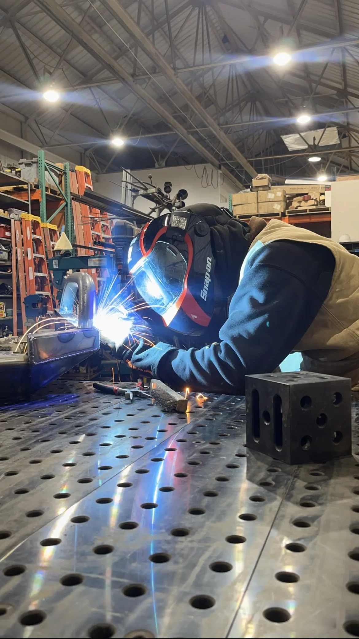 A person welding metal in a workshop, wearing a protective helmet and gloves, with sparks and bright light from the welding process, surrounded by industrial shelves and equipment.