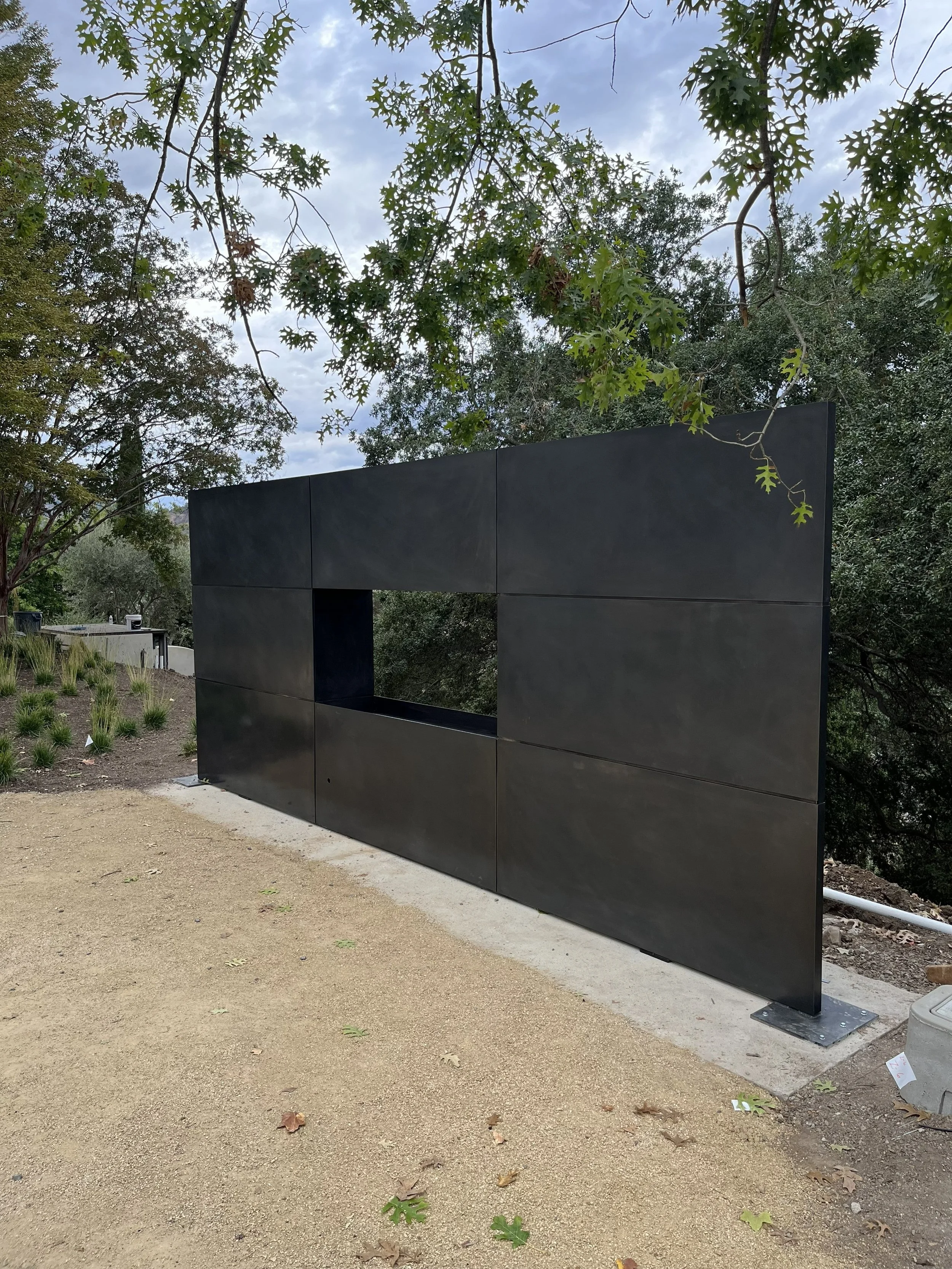 A modern black metal sculpture with a rectangular opening, situated outdoors among trees and a cloudy sky.
