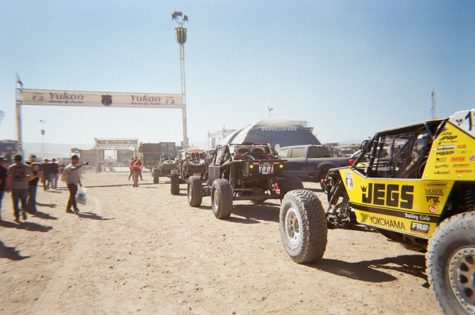 Off-road racing vehicles parked on a dusty race track with people walking around, a large banner reading Yukon, and a clear blue sky.