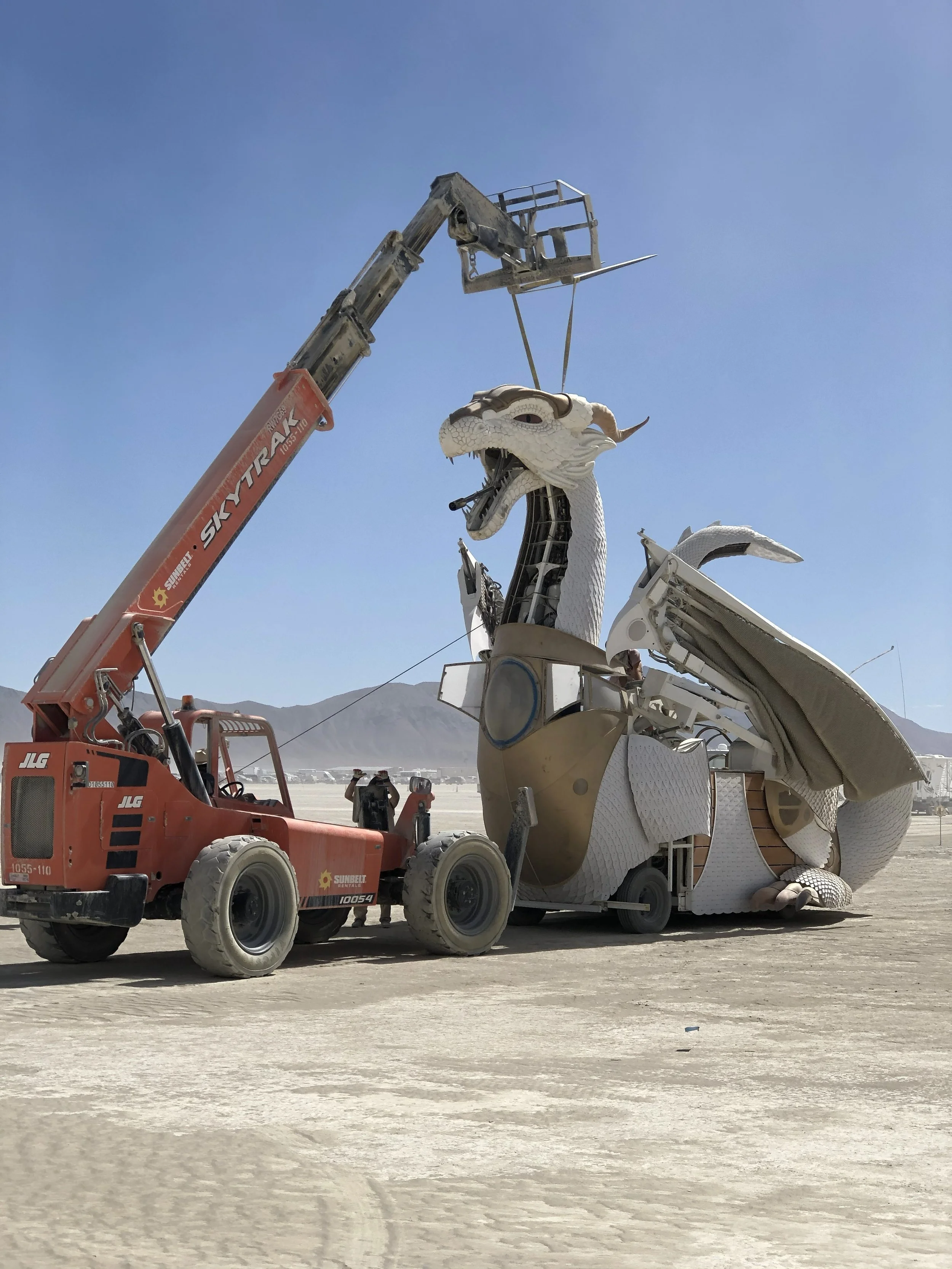 A large white dragon sculpture being assembled with a crane at a desert festival, with people in the background and mountains in the distance.