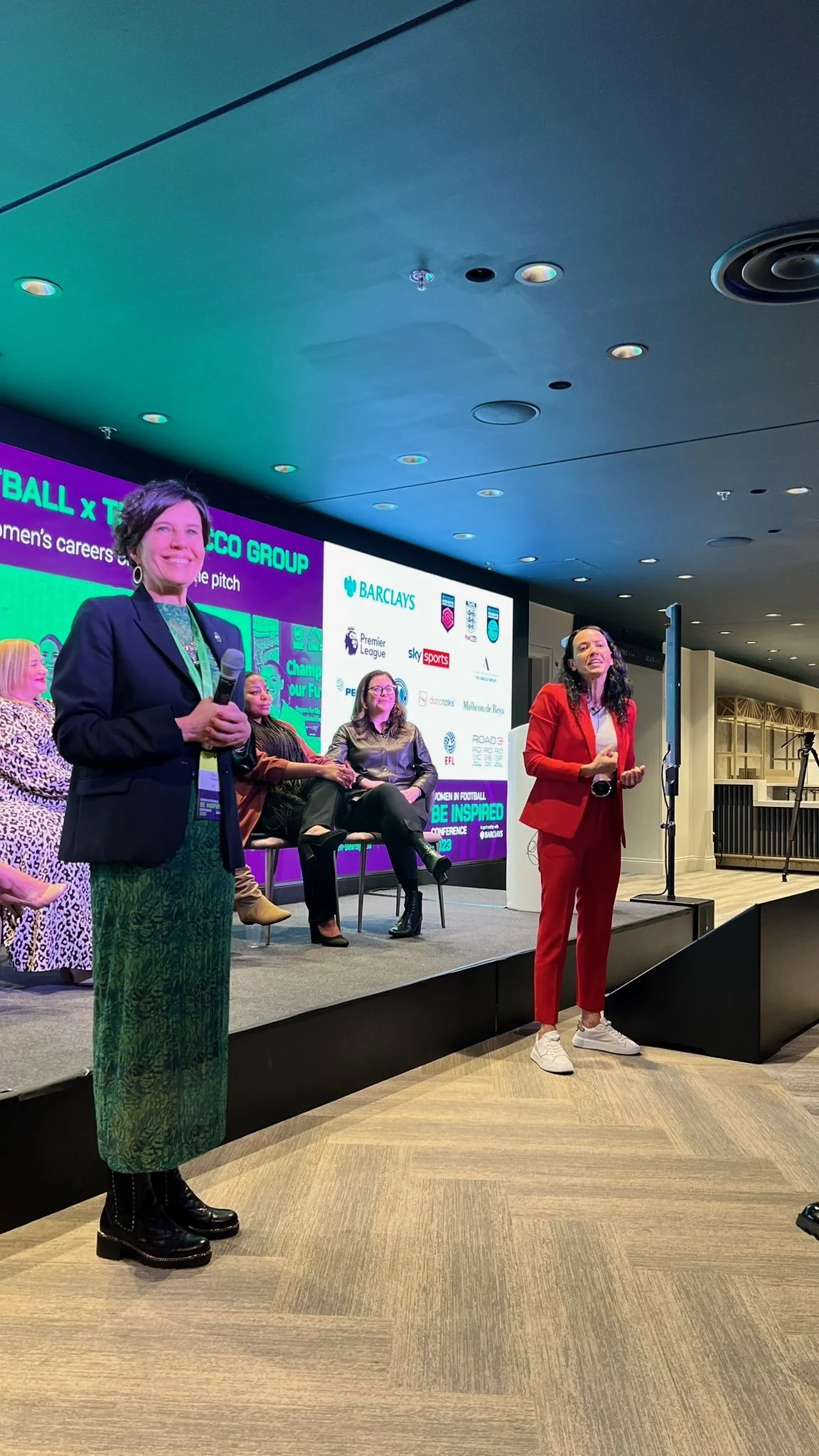 Women on stage at a conference, with one woman standing and holding a microphone, others sitting and listening, a large screen behind them displaying sponsor logos and conference details.