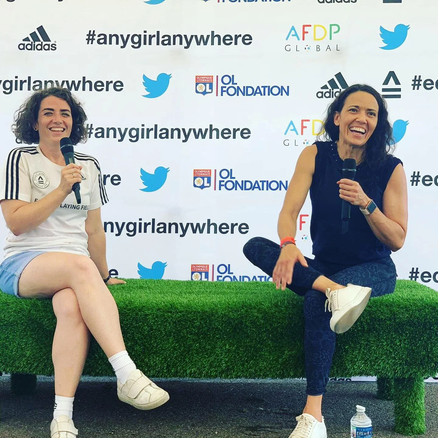 Two women sitting on a green grass-like platform, each holding a microphone. They are smiling and engaging with an audience against a backdrop with hashtags and logos, including #angirlanywhere, Twitter, Adidas, and OL Fondation.