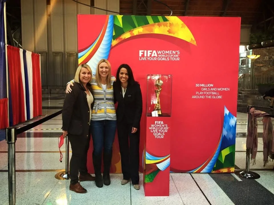 Three women standing in front of a FIFA Women's World Cup 2016 display at an indoor venue, smiling for a photo.